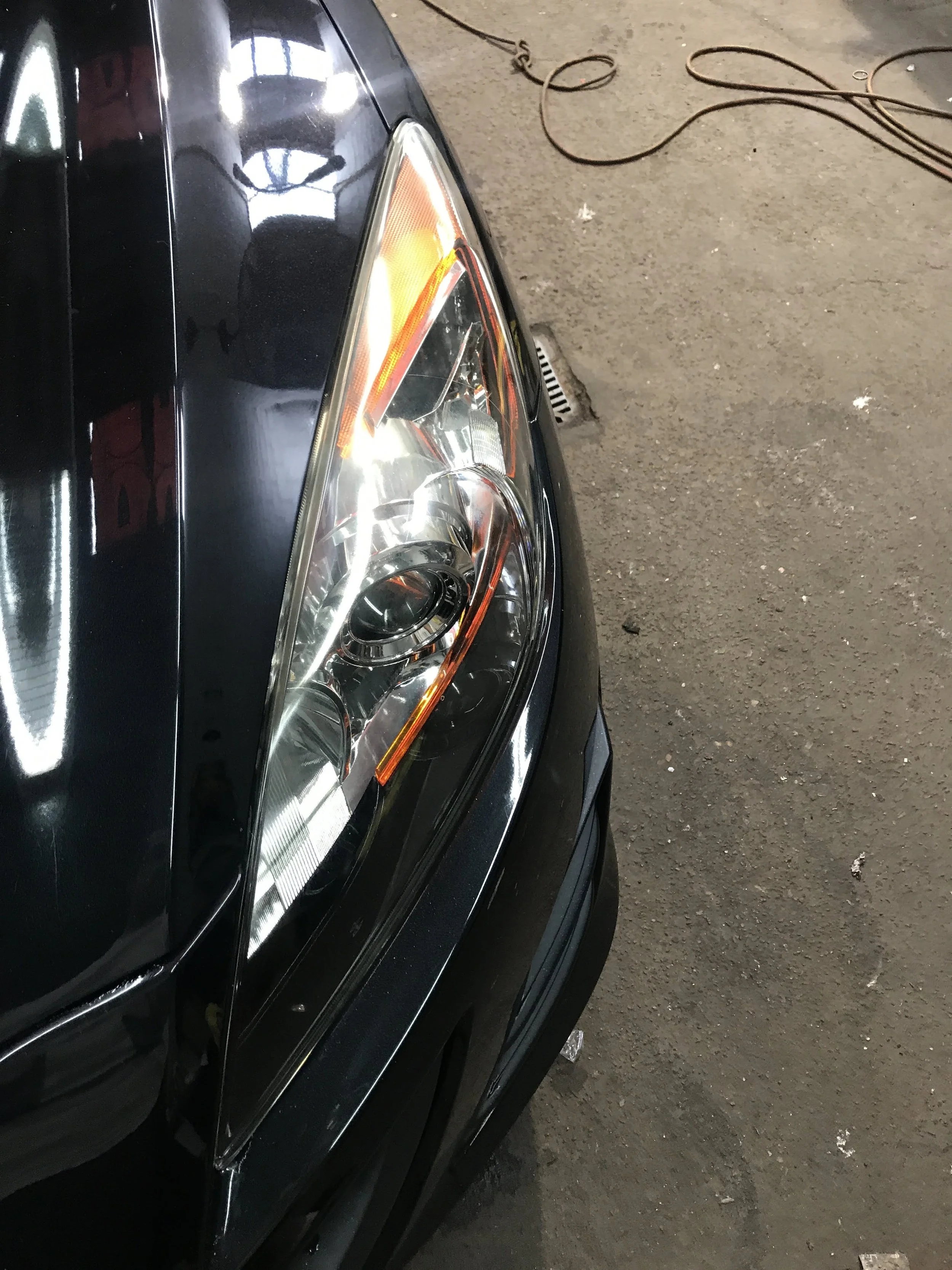 Close-up of the front part of a black car with a large headlight, parked on a concrete surface with a drain and electrical cords nearby.