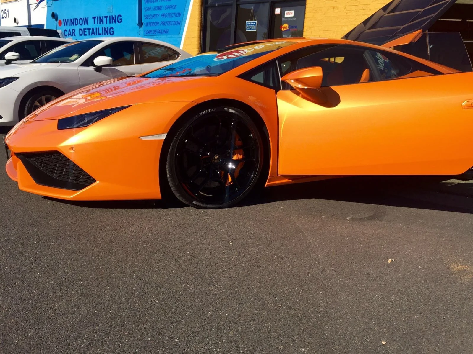 An orange Lamborghini sports car with black wheels parked in a lot, with white and silver cars behind it and a building with a blue sign advertising car detailing in the background.
