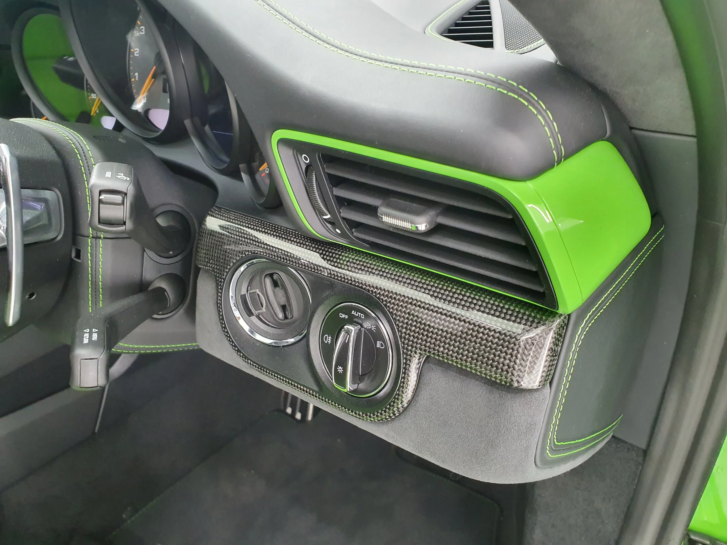 Close-up of a car's interior dashboard, featuring green accents, air vents, a round control dial, and a carbon fiber center console.