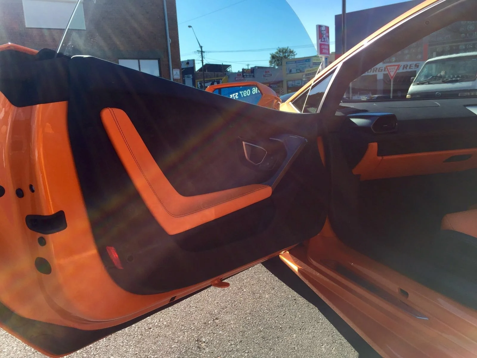 The interior of an orange sports car, showing the driver's side door open, with orange and black seats, and a city street with buildings and cars visible through the windshield.