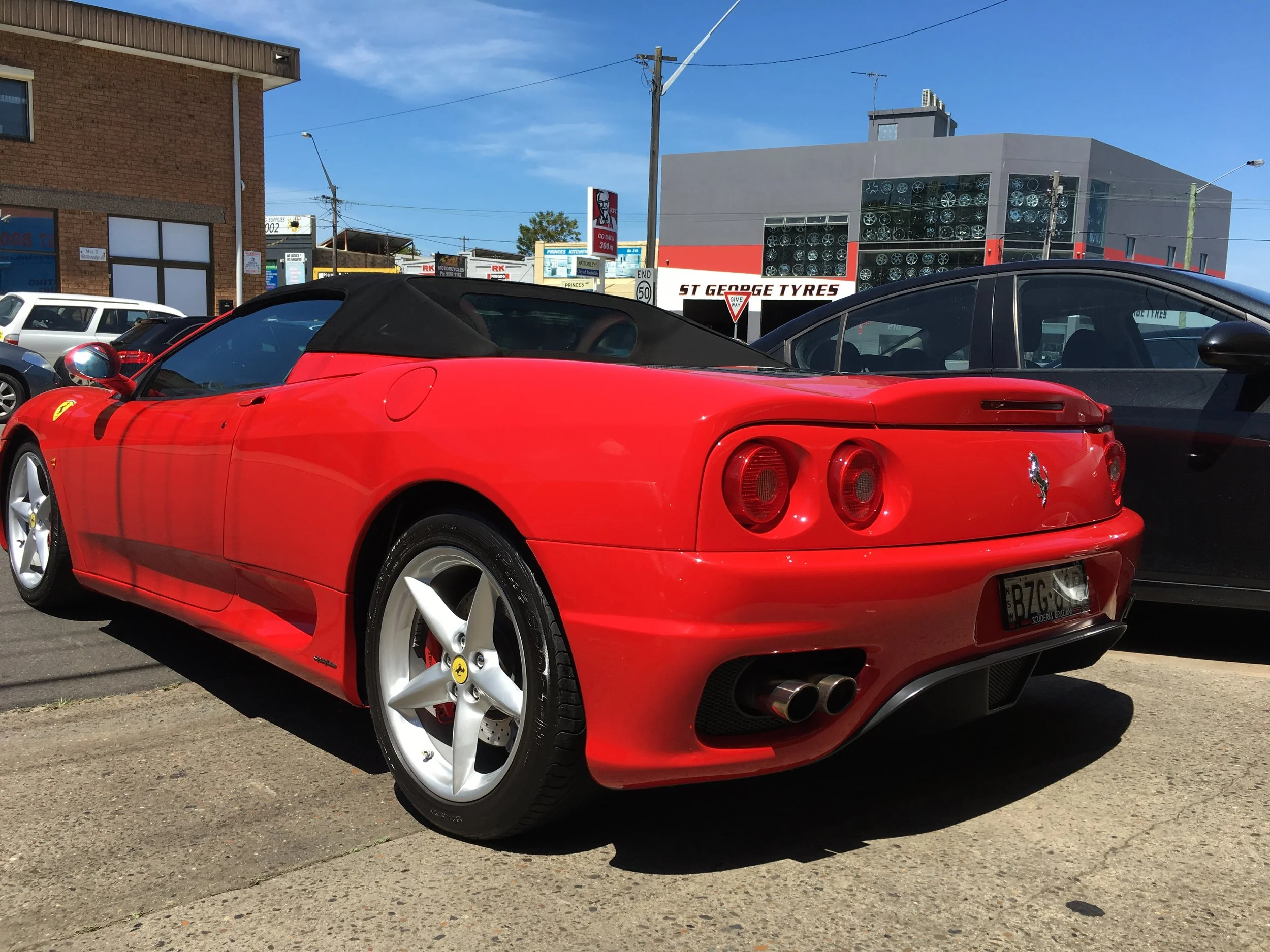 Red Ferrari convertible parked on the street with a row of other cars and buildings in the background. The Ferrari has a black soft top and white wheels.