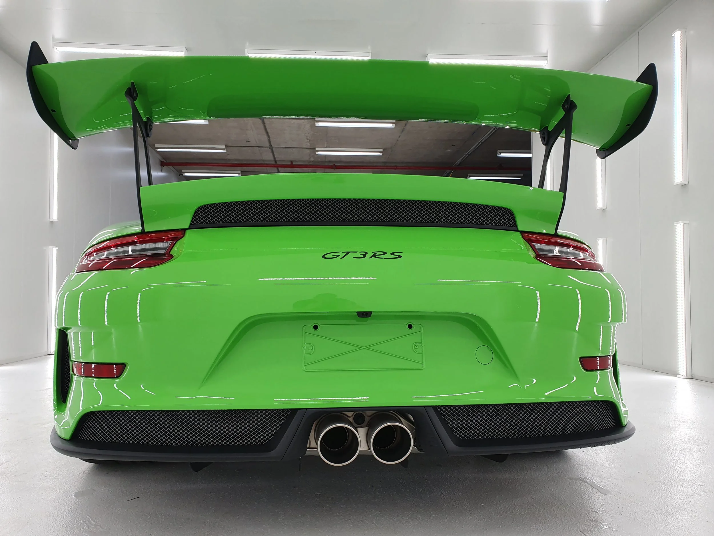 Rear view of a bright green sports car with a large rear wing, quad exhaust pipes, and the model name 'GT3 RS' on the back, parked in an indoor garage with white walls and bright lighting.