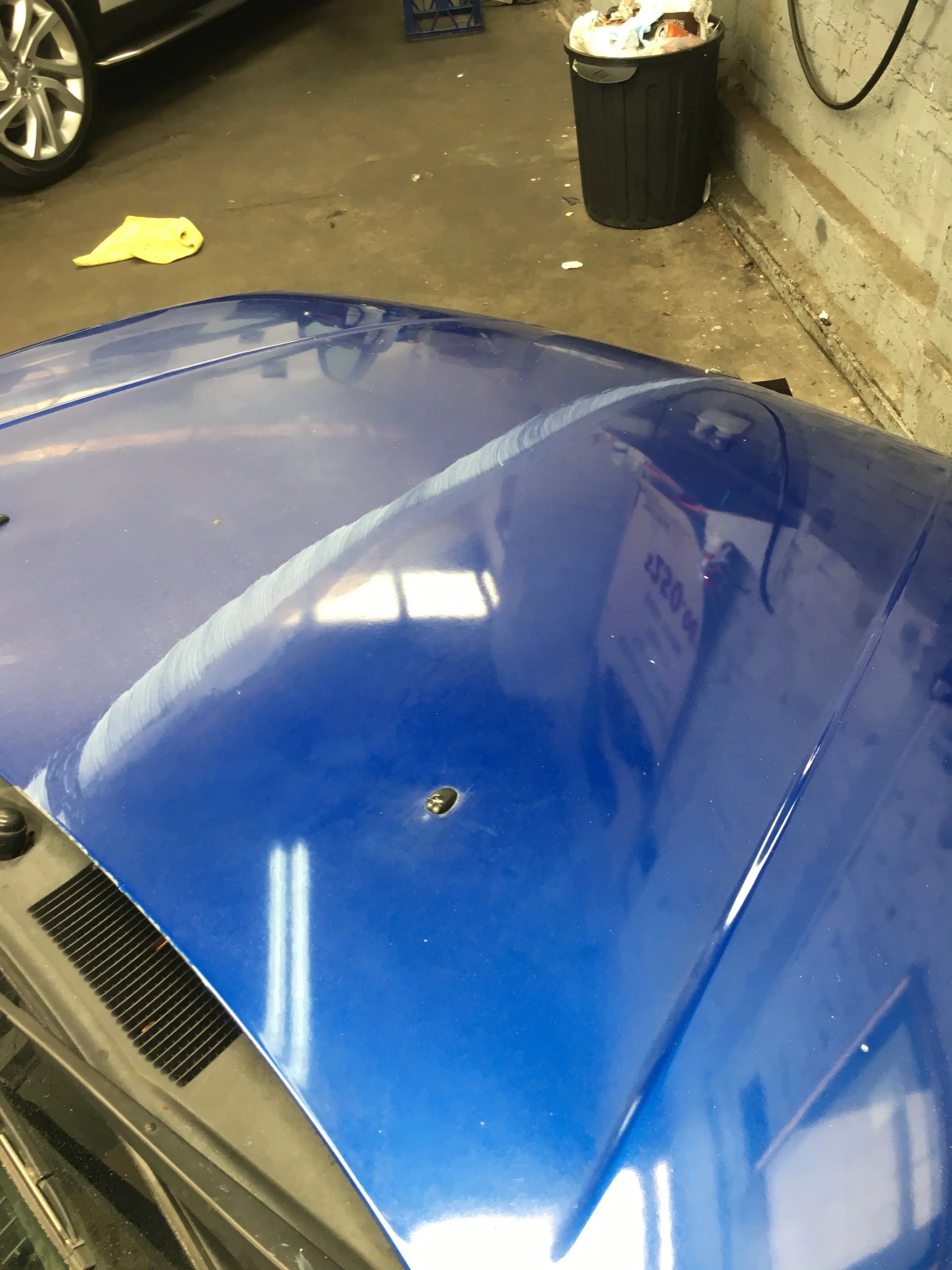 Close-up of the blue hood of a car parked in a garage, with part of a black car and a yellow cloth on the ground nearby, and a trash can filled with trash in the background.