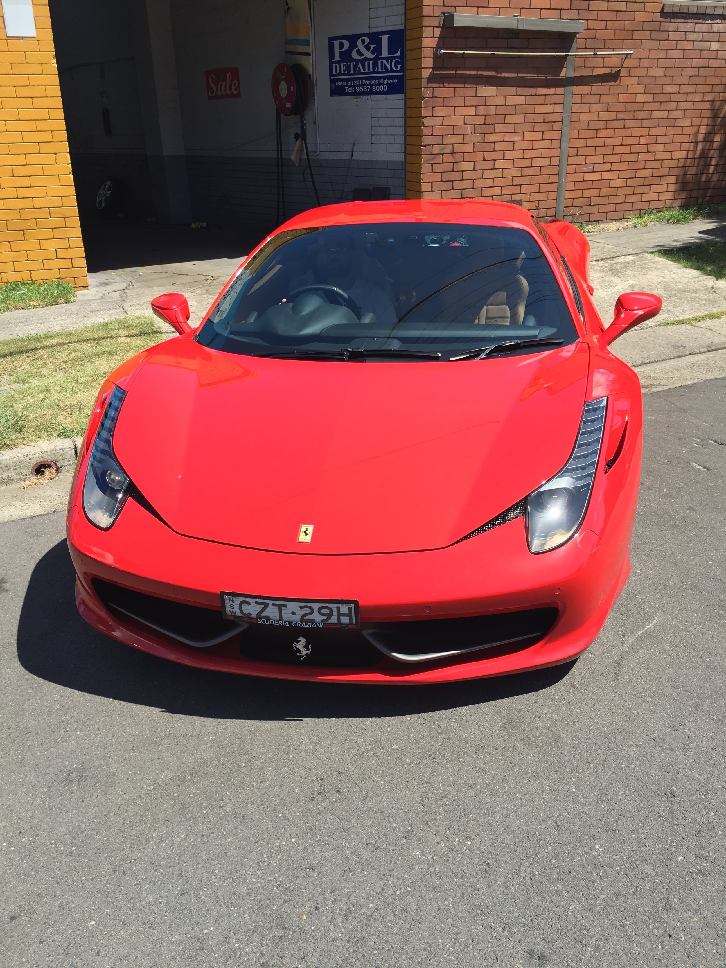 Red Ferrari sports car parked on the street, with a car dealership and brick building in the background.