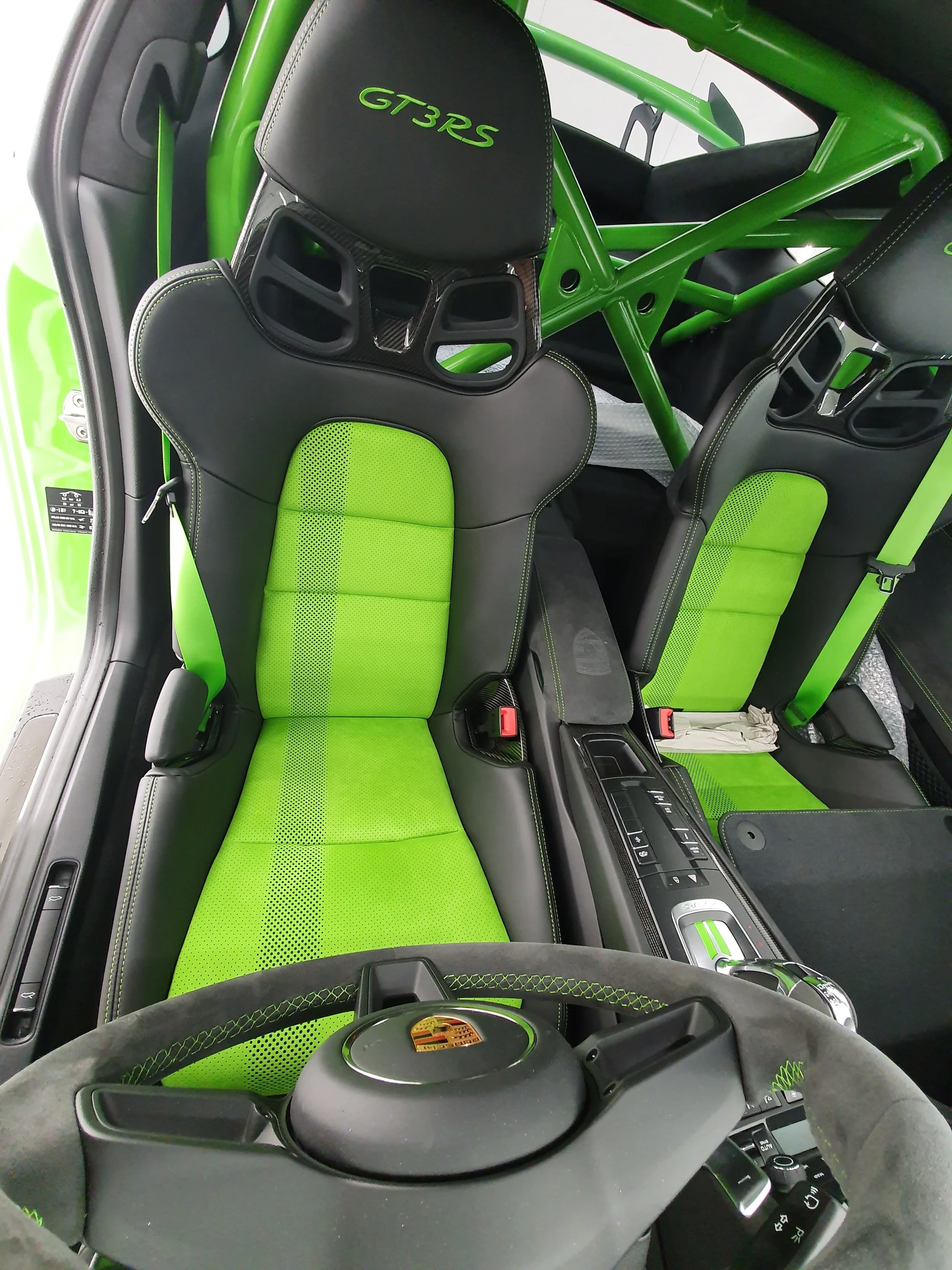 Interior of a green Porsche race car, featuring black and green racing seats with the Porsche logo on the steering wheel, and a roll cage with 'GT3RS' embroidered on the headrest.