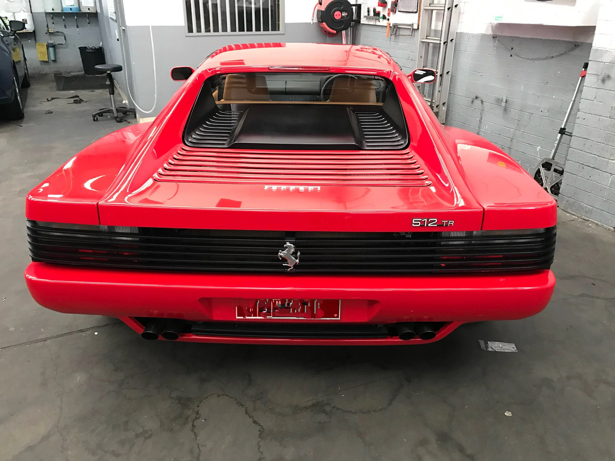 Rear view of a red Ferrari 512 TR sports car parked inside a garage with gray brick walls, tools on the wall, a hanging hose reel, and garage equipment.