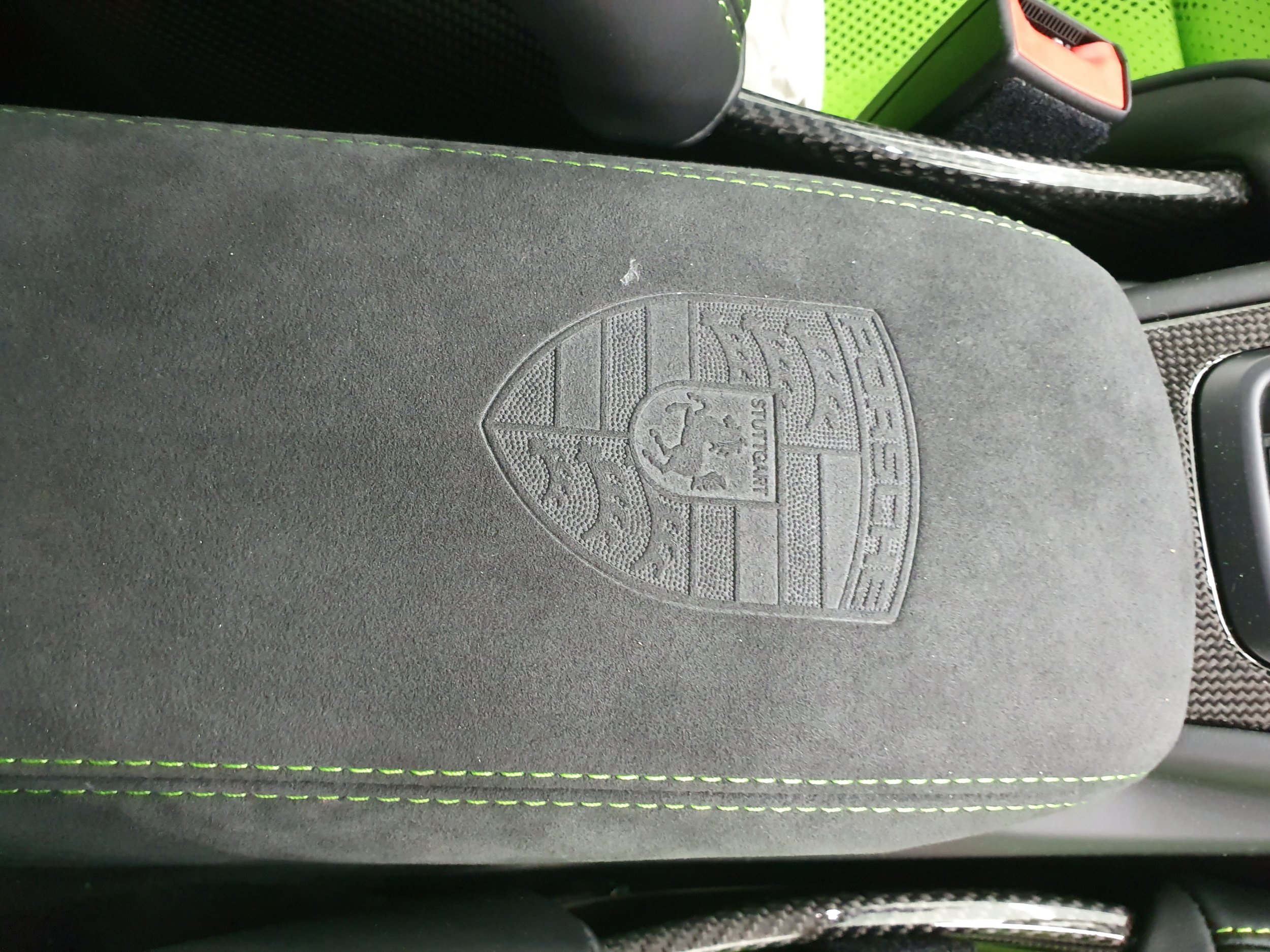 Close-up of a car seat with the Porsche emblem embossed on the headrest, featuring black upholstery with green contrast stitching, and other interior elements visible.
