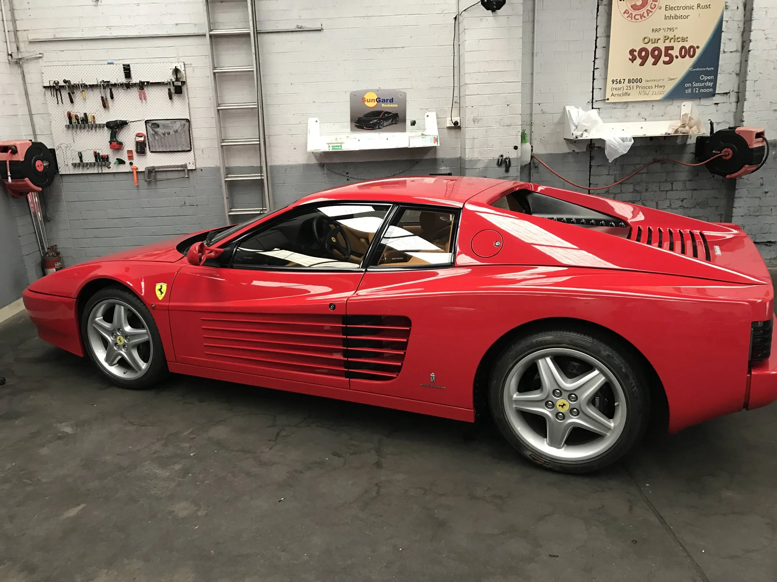 Colorful workshop with a red Ferrari sports car parked indoors. The background has tools hanging on a pegboard, a folded ladder, and posters on the white brick walls, including one advertising electronic rust inhibitor for $995. The floor is dark con