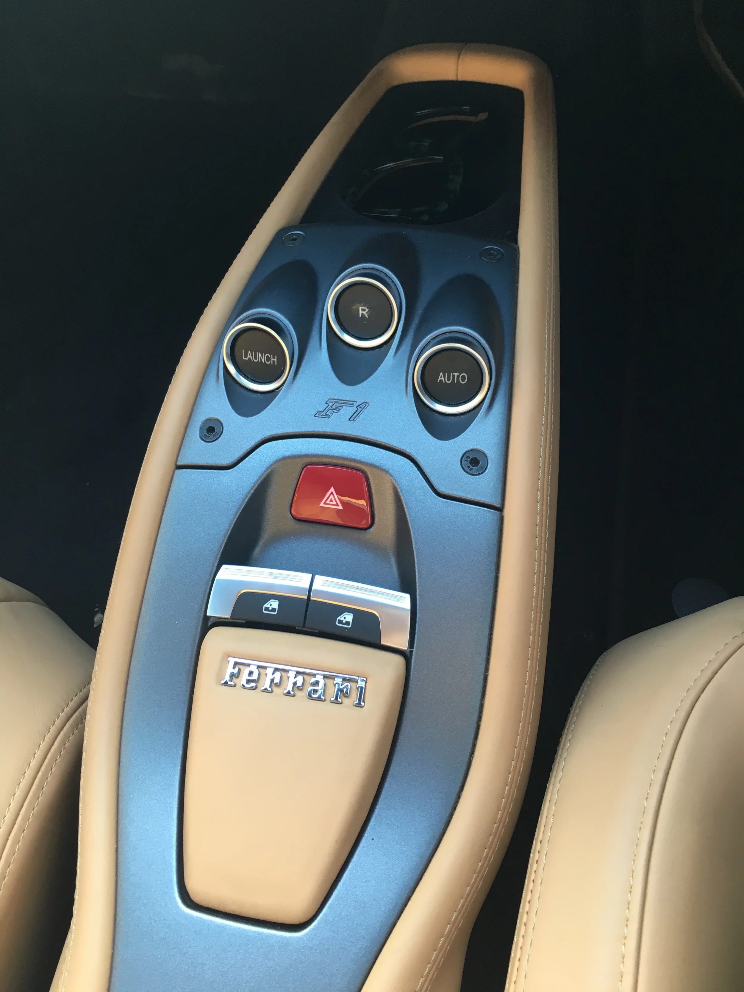 Center console of a Ferrari car with buttons labeled R, AUTO, and LAUNCH, beige leather surrounding the console, and a Ferrari logo at the bottom of the console.