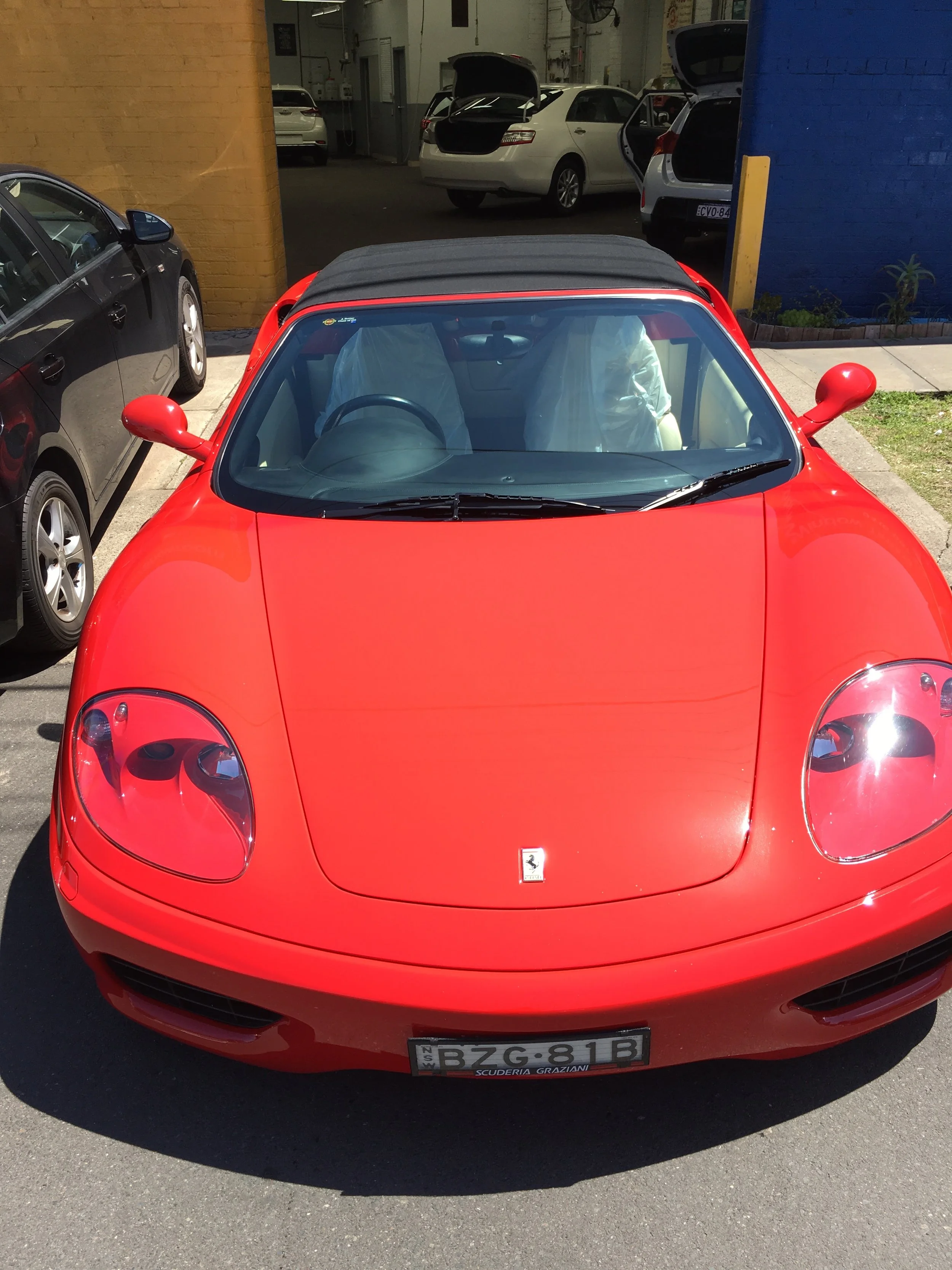 A red Ferrari sports car parked outdoors, with the front view showing the Ferrari logo on the hood, headlight details, and license plate reading BZG-81B.