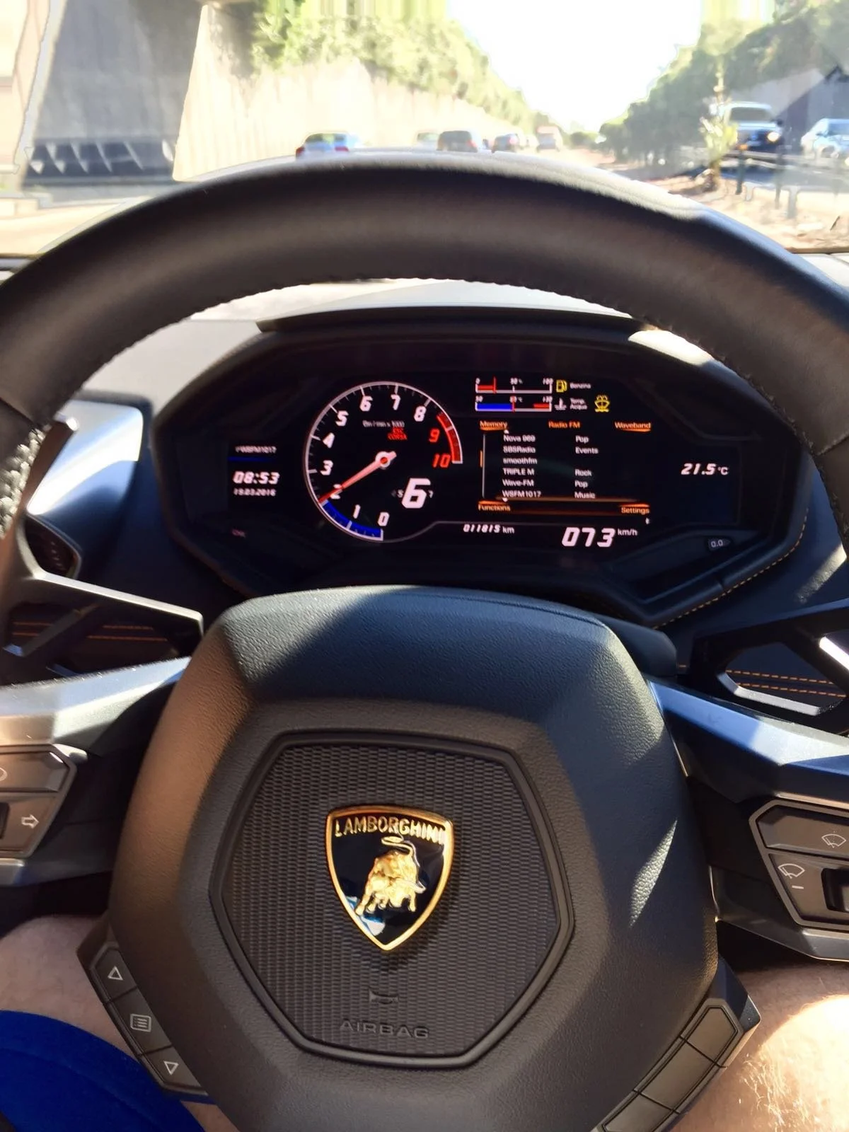 The photo shows the interior dashboard and steering wheel of a Lamborghini car, with a digital display indicating speed, temperature, and other vehicle information. The steering wheel prominently features the Lamborghini logo.