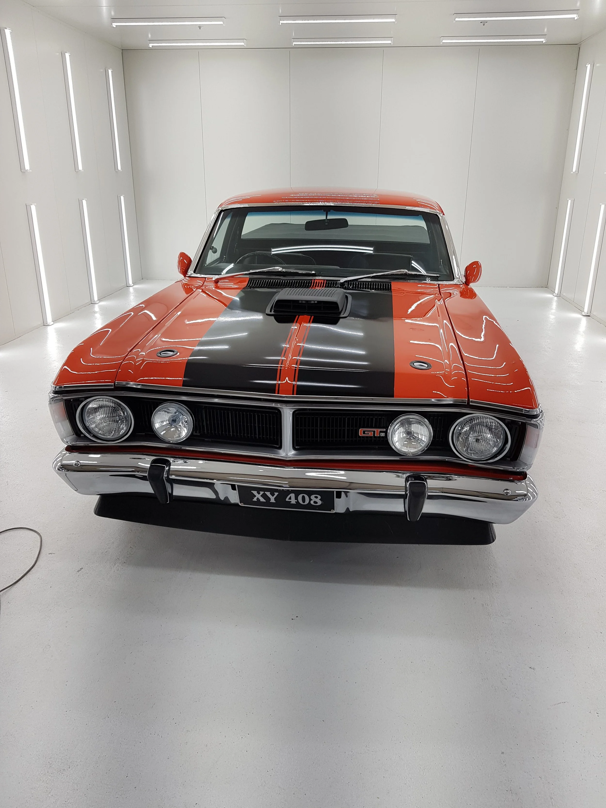 A vintage red and black Ford XY 408 muscle car with a hood scoop, parked in a white enclosed room.