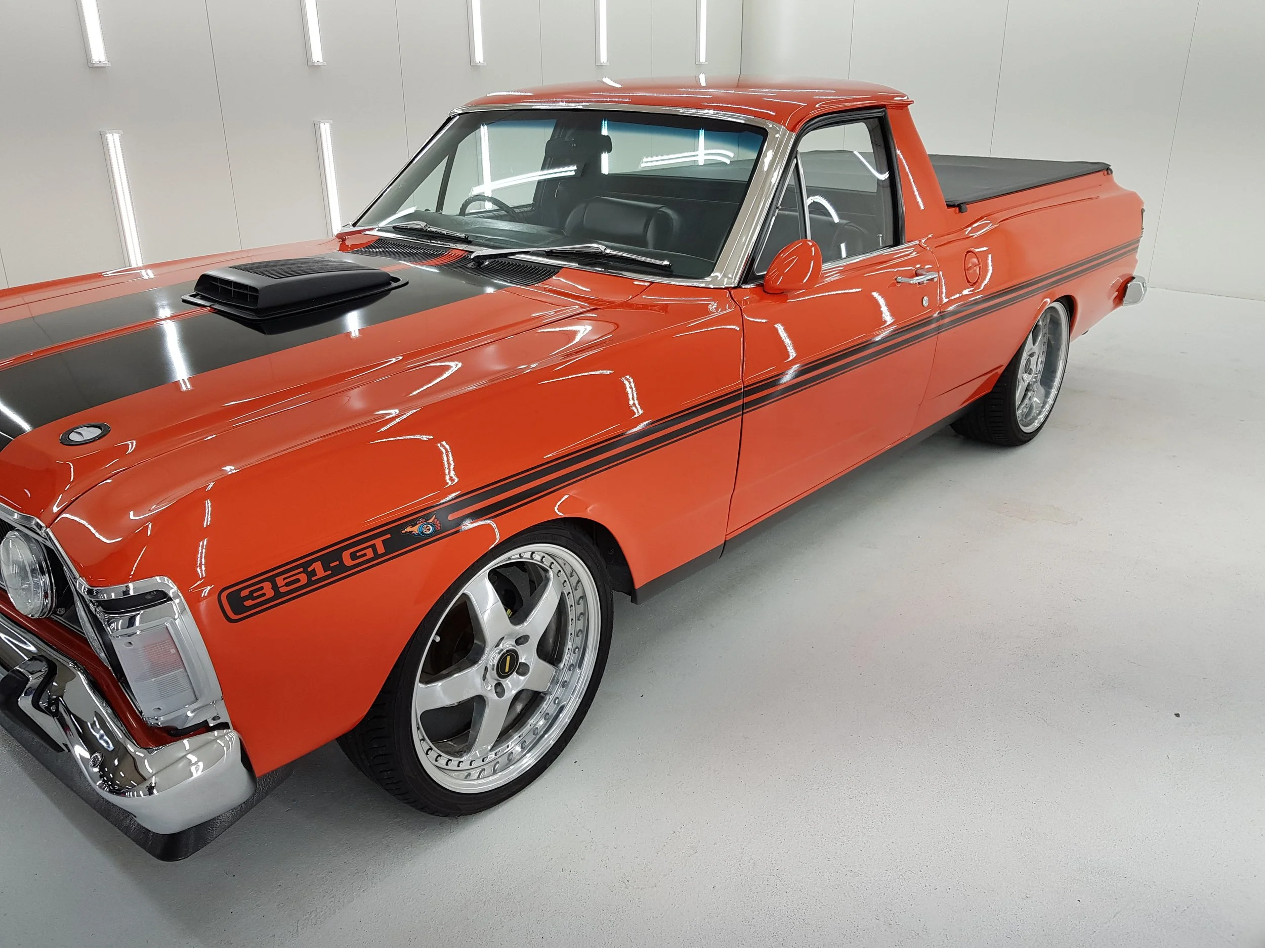 A red classic car with black racing stripes and a matte black hood scoop, parked in a bright white room with vertical lighting.