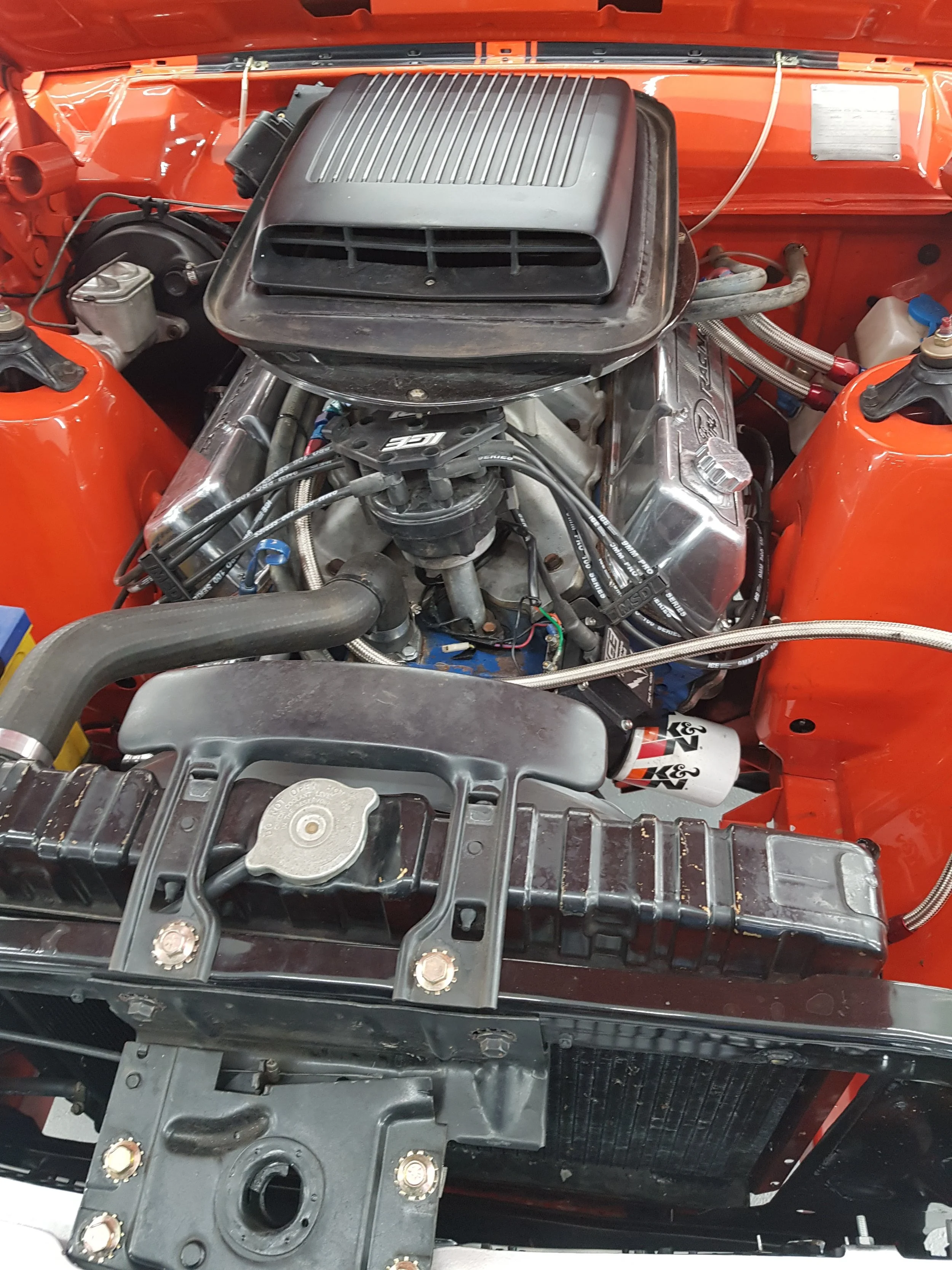 The image shows the engine bay of a racing vehicle with an air intake system, various hoses, and metallic components, along with other automotive parts.