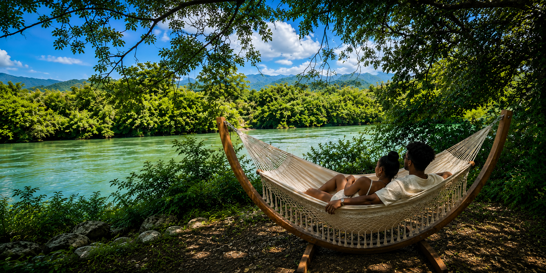 Deux personnes allongées dans un hamac en bois au bord d'une rivière entourée de verdure dense et de montagnes en arrière-plan, sous un ciel bleu avec quelques nuages.