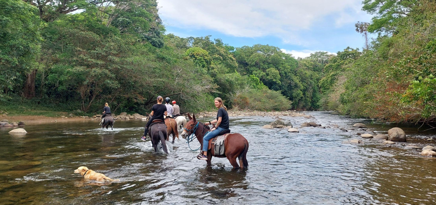Groupe de personnes à cheval traversant une rivière dans une forêt verdoyante sous un ciel partiellement nuageux.