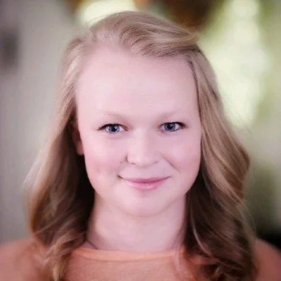 A young woman with blonde hair smiling at the camera, with a blurred outdoor background.