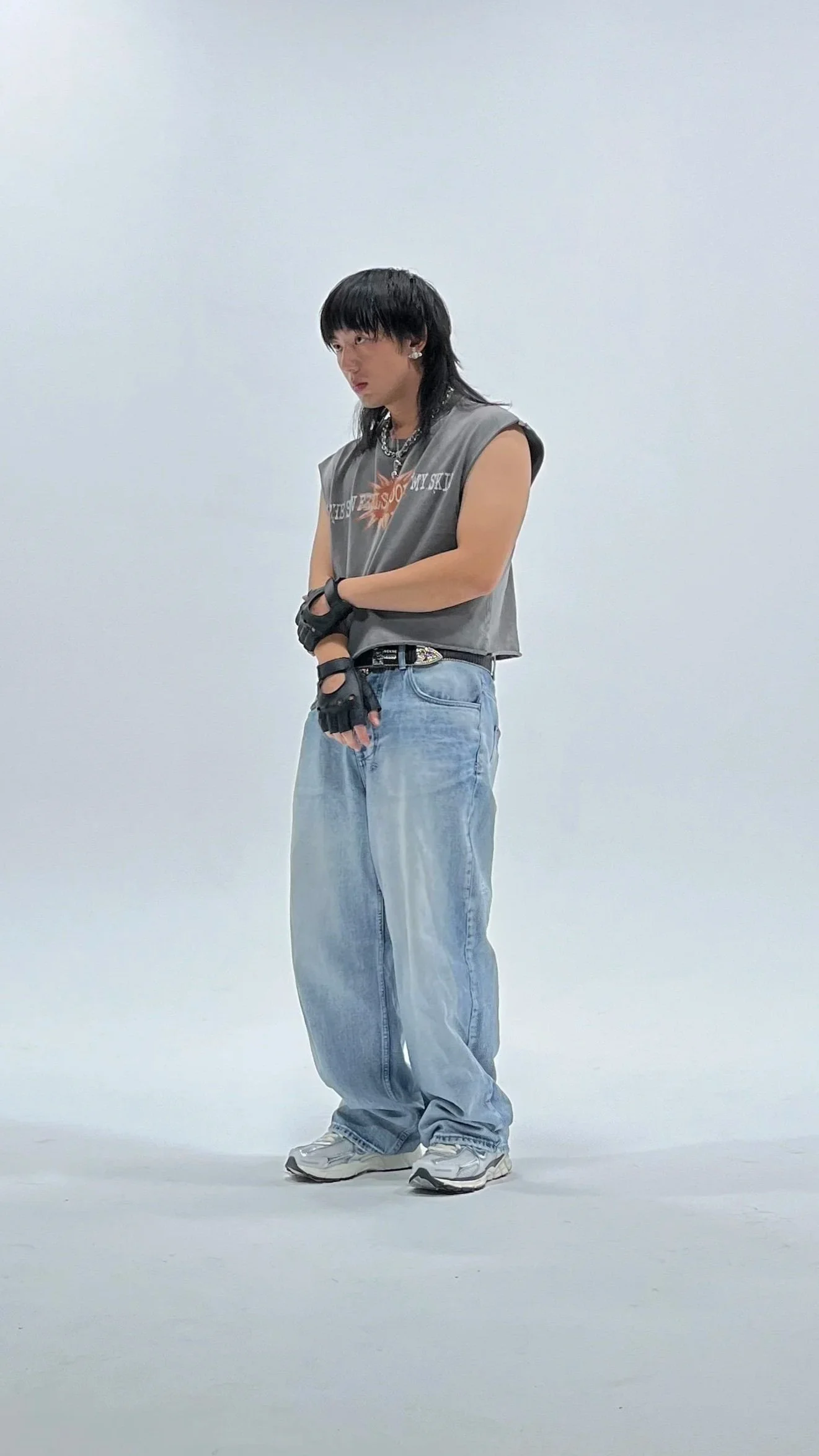 A person with dark hair and bangs, wearing a sleeveless top, baggy jeans, sneakers, and gloves, standing in a white cyc wall studio with black ceiling panels.