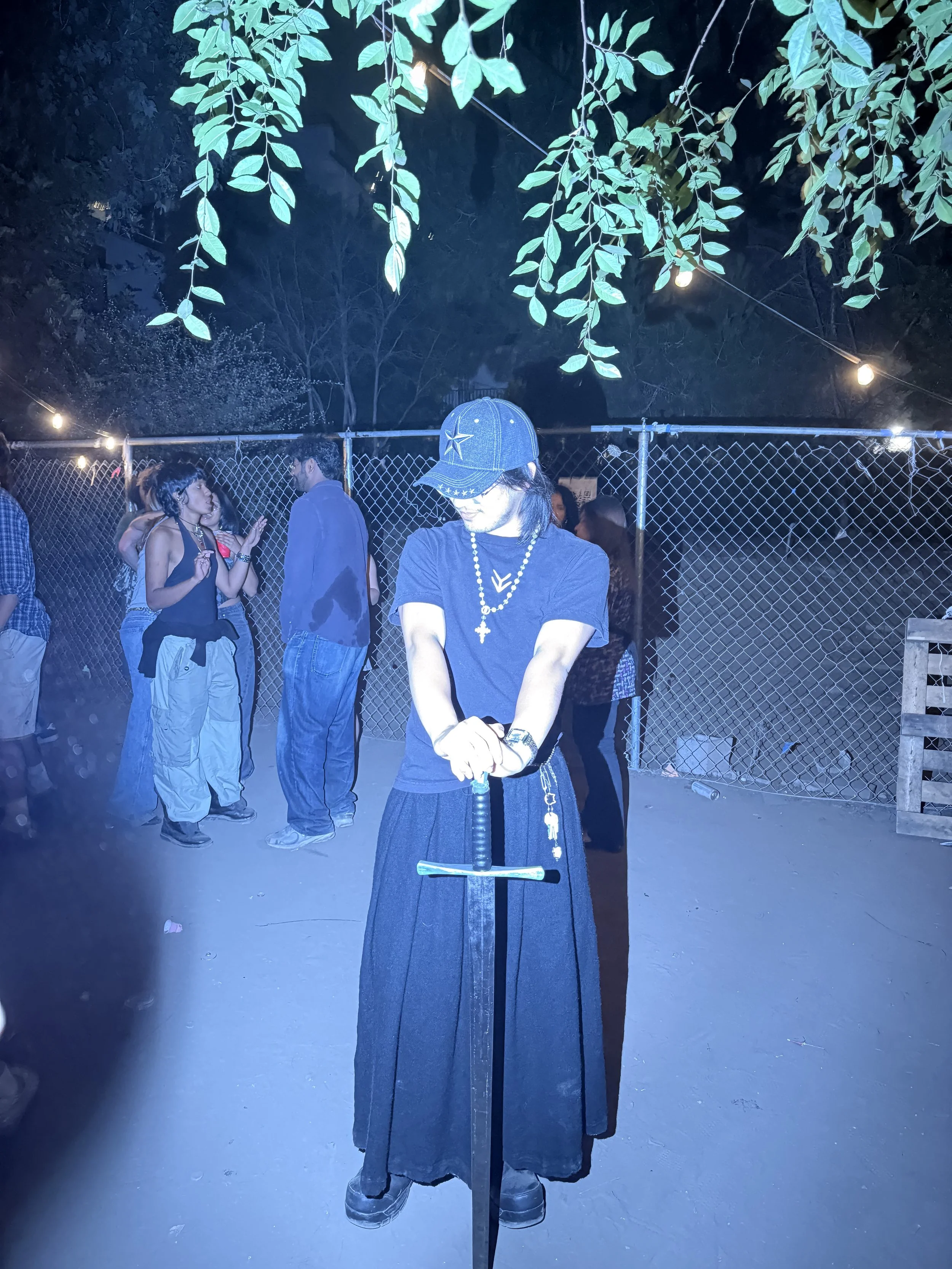 A person dressed in gothic attire, wearing a black hat with white stars, a black dress, and jewelry, standing at night with their arms crossed in front of a sword plunged into the ground. In the background, several people are talking and socializing 