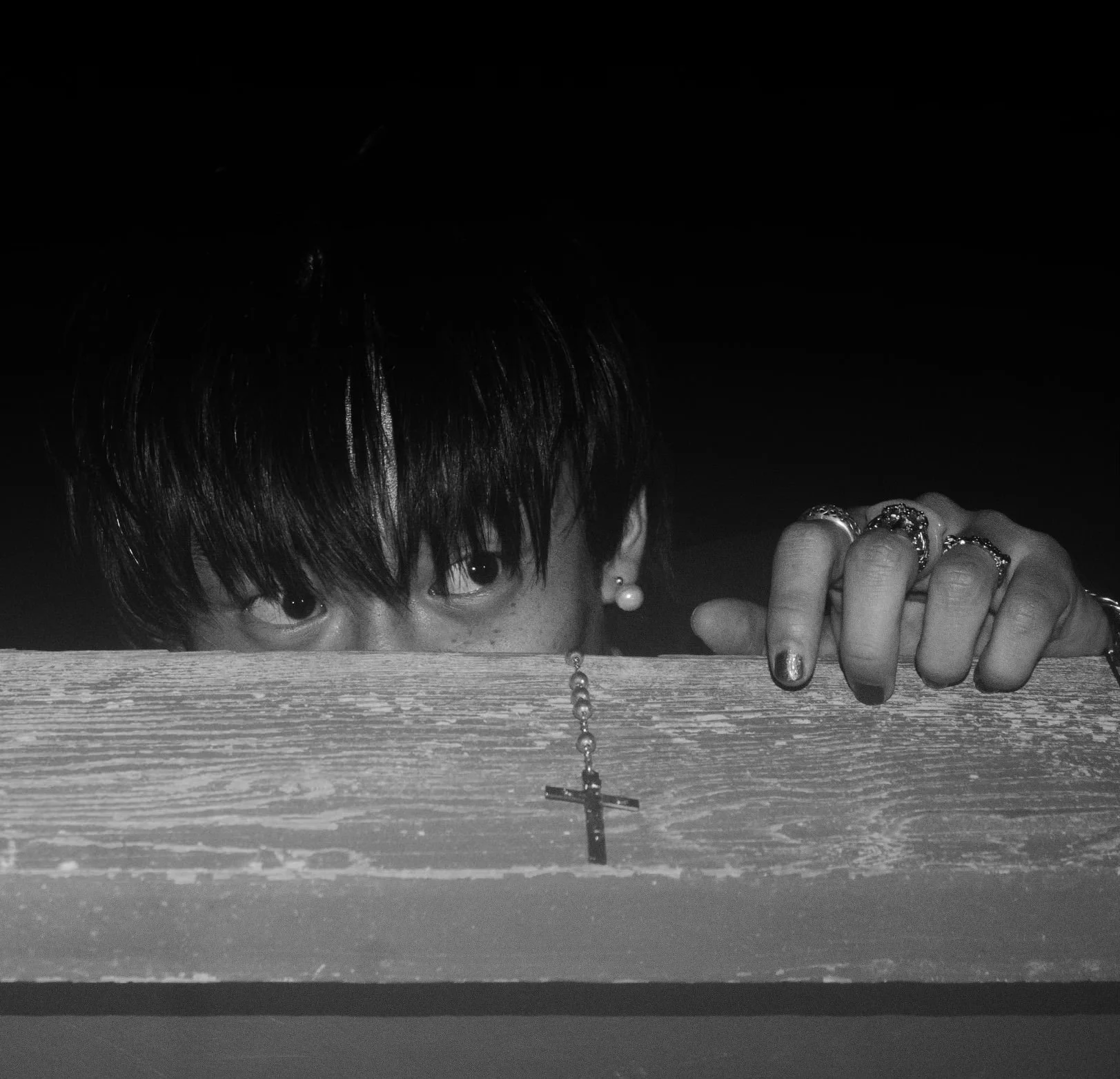A person with dark hair and earrings peeking over a wooden surface, holding a cross necklace with fingers adorned with rings, against a dark background.