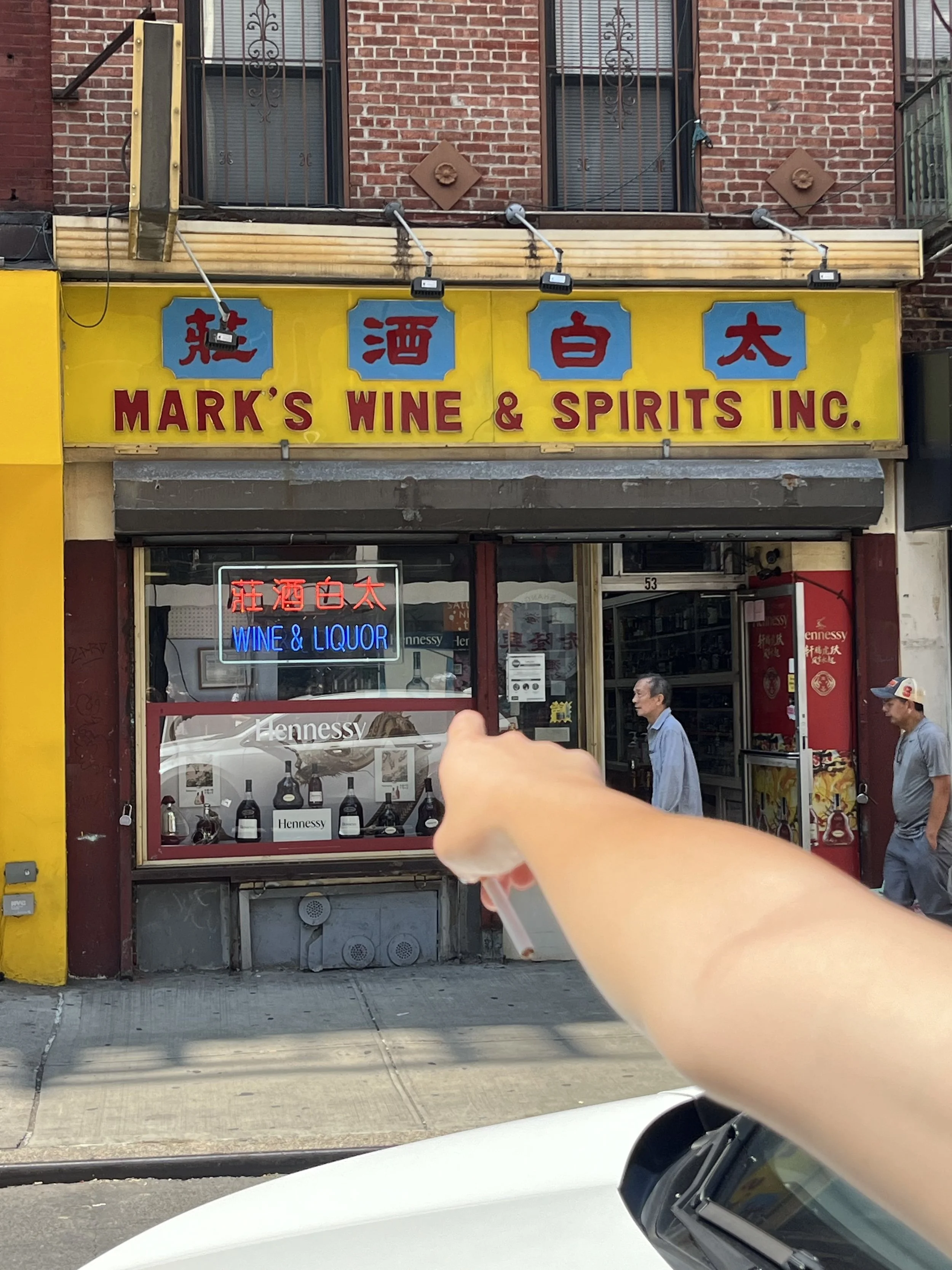 Street scene with a storefront sign for 'Mark's Wine & Spirits Inc.', displaying Chinese characters, a neon sign for wine and liquor, and pedestrians passing by.