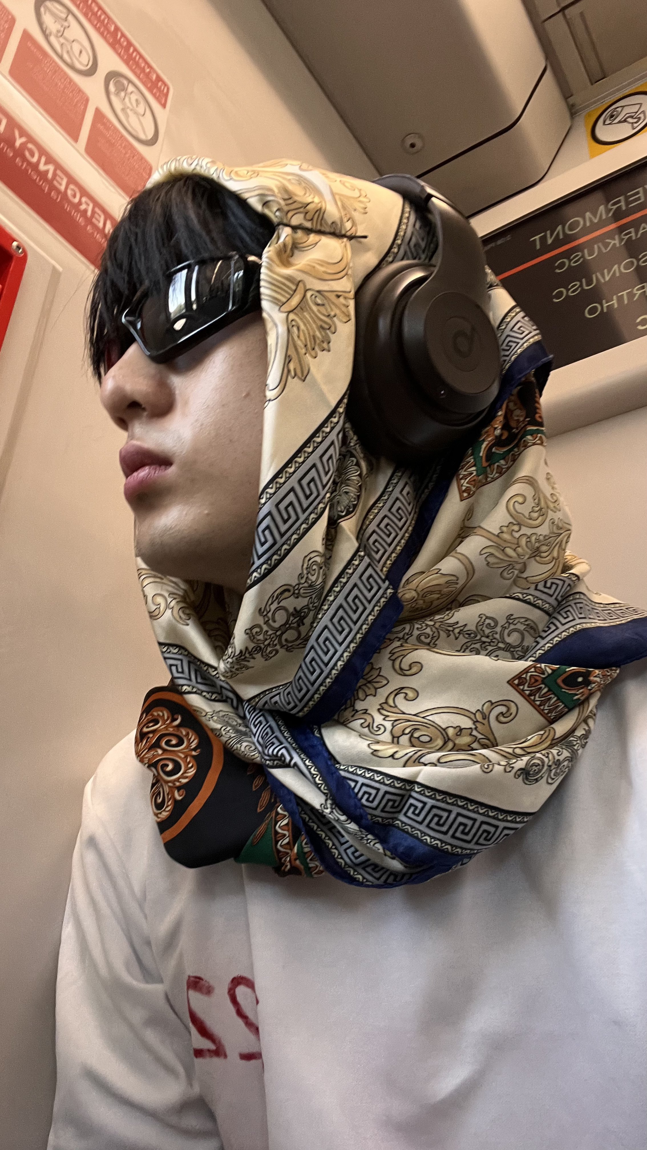 tu. wearing black sunglasses, a patterned silk scarf around his head, and a white sweatshirt with red lettering on the sleeve, sitting on an airplane or train, with safety and warning signs visible in the background.