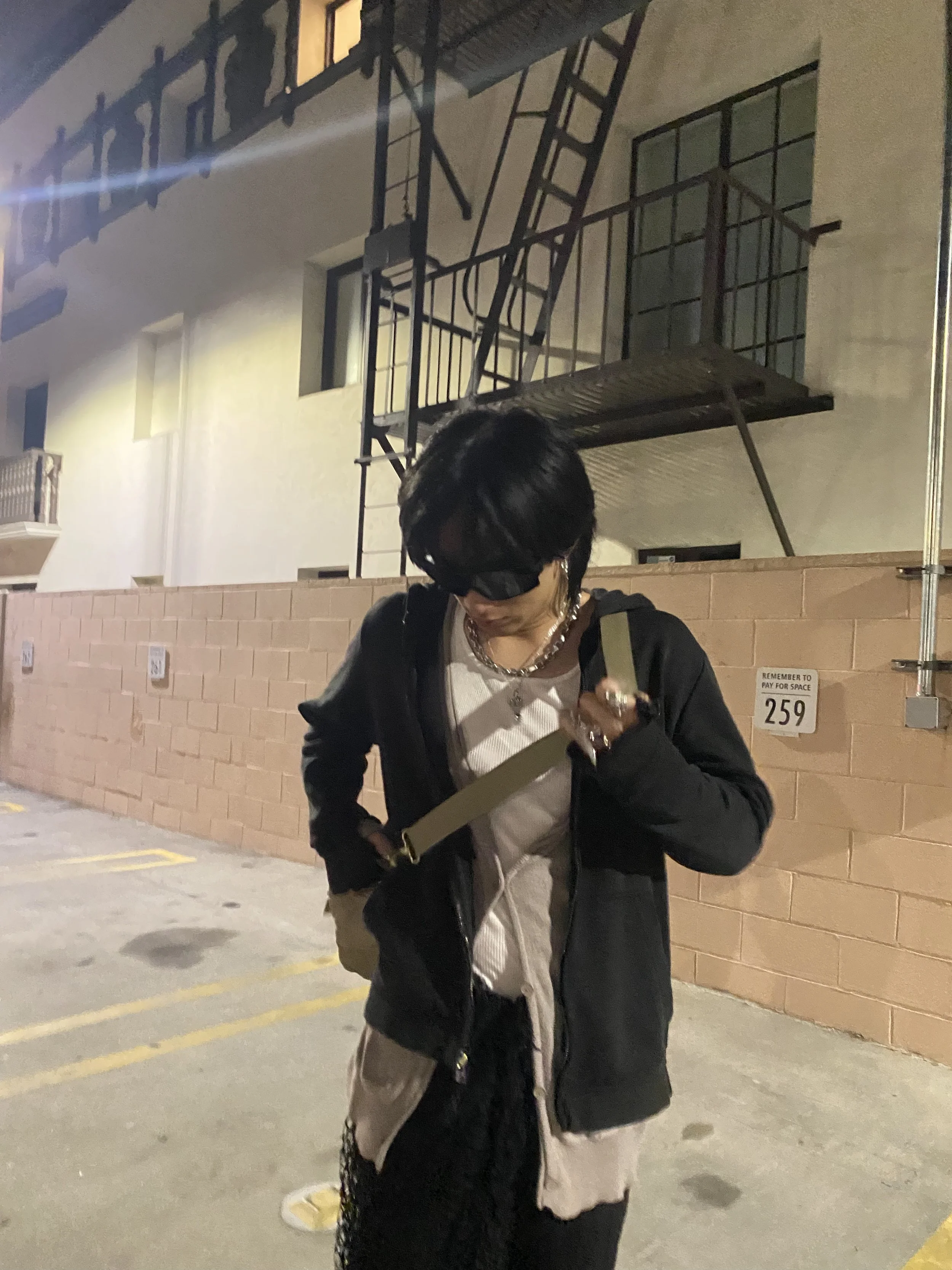 tu. with black jacket, holding a phone, standing in a parking lot at night with a brick wall and apartment windows behind them.