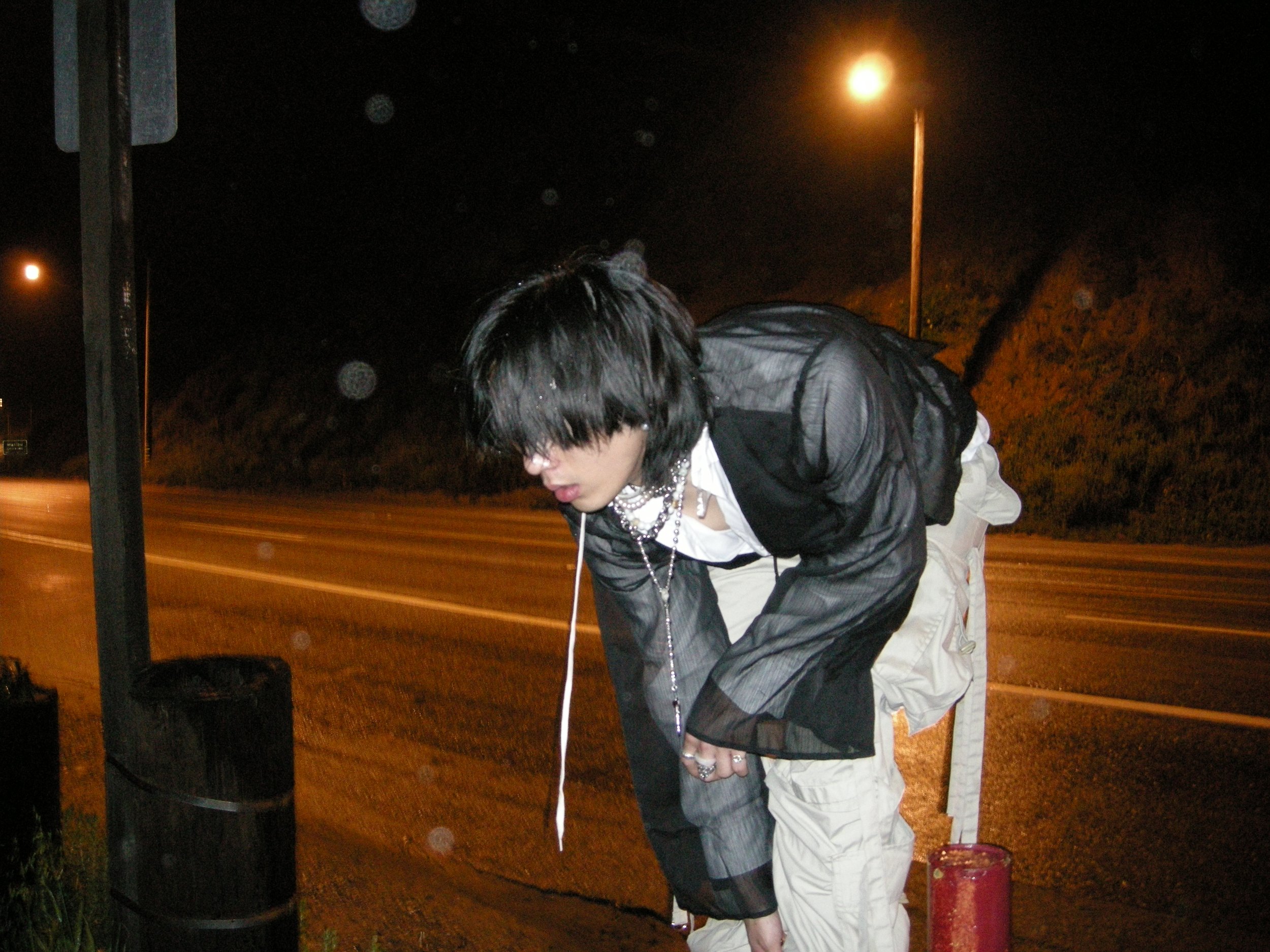 tu. with layered necklaces, wearing a sheer black jacket over a white shirt, is leaning down outdoors at night near a street lamp and a trash can, with raindrops visible on the camera lens.