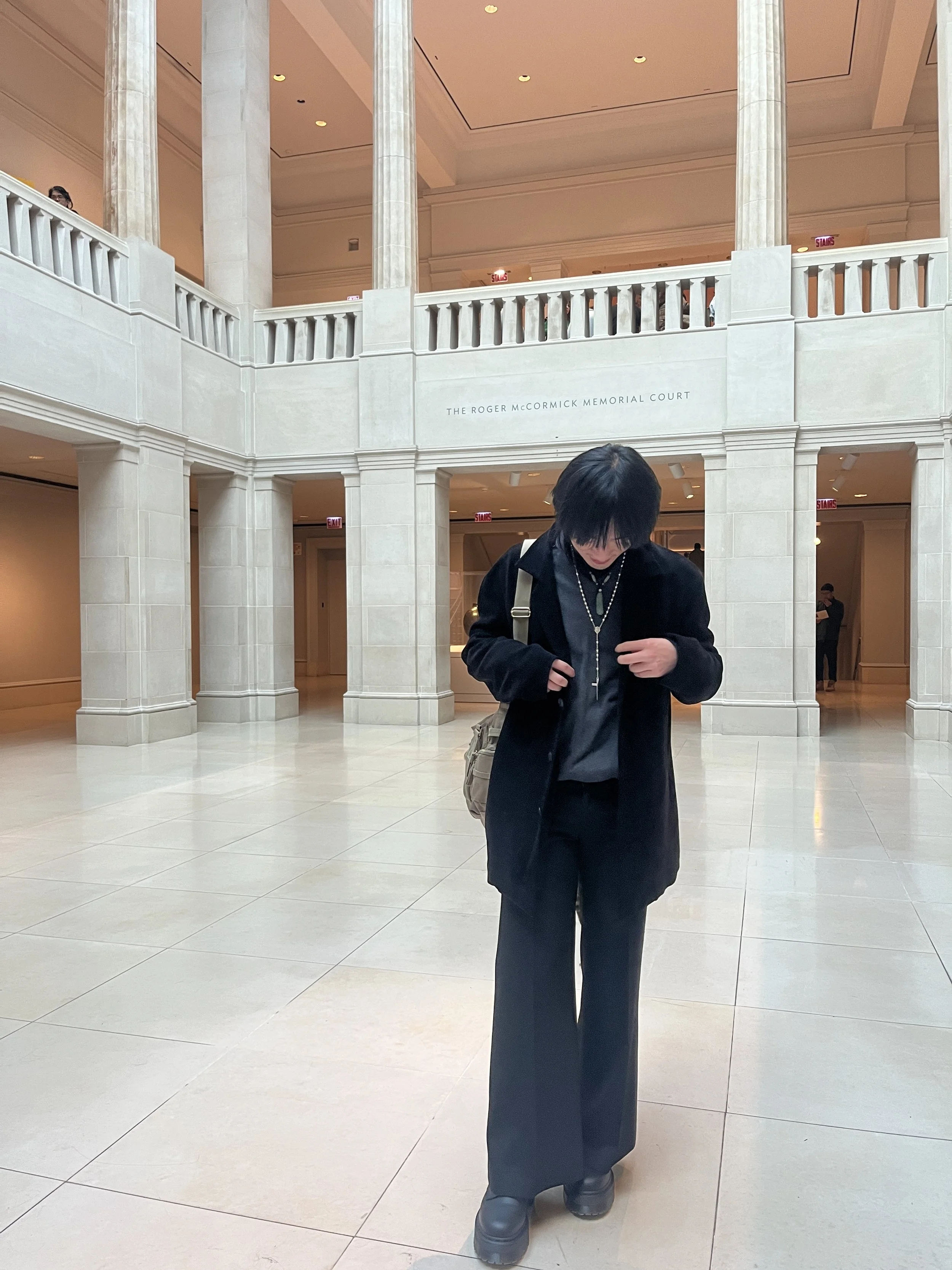 tu. wearing a black coat and black pants, stands inside The Roger McCormick Memorial Court, looking down. The interior features beige walls, large columns, and a mezzanine level with a railing. There are signs indicating stairs and exits.