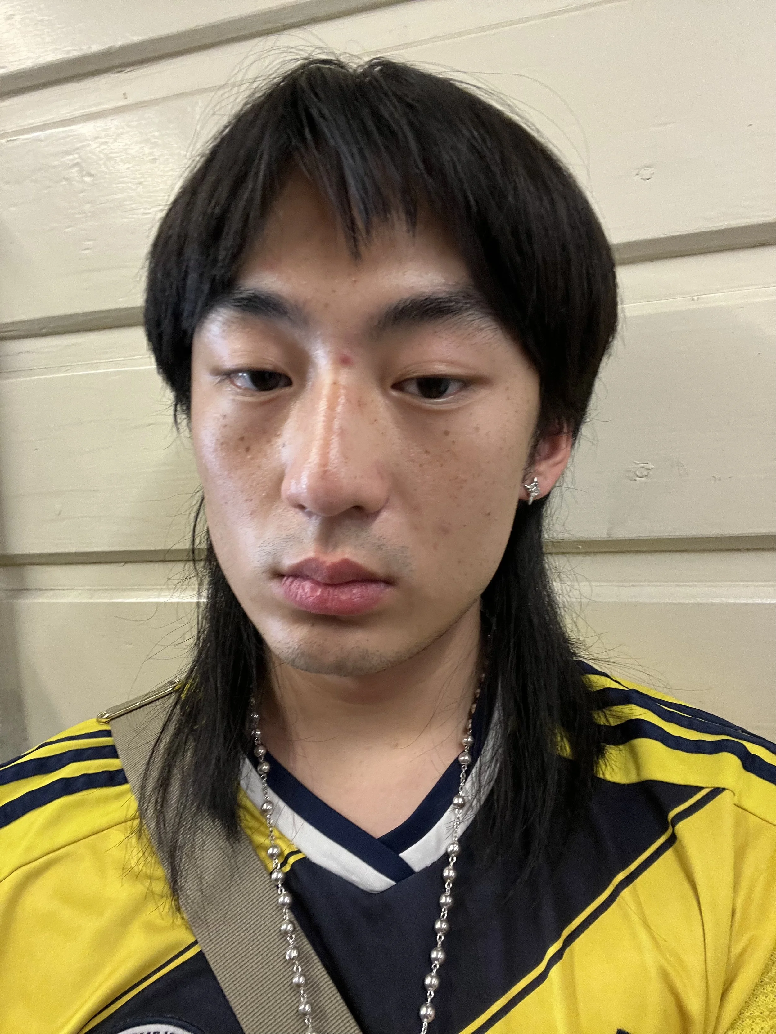 tu. wearing a yellow jacket with black stripes, a navy blue and white shirt, a gray messenger bag, and a silver beaded necklace, standing against a beige wall.