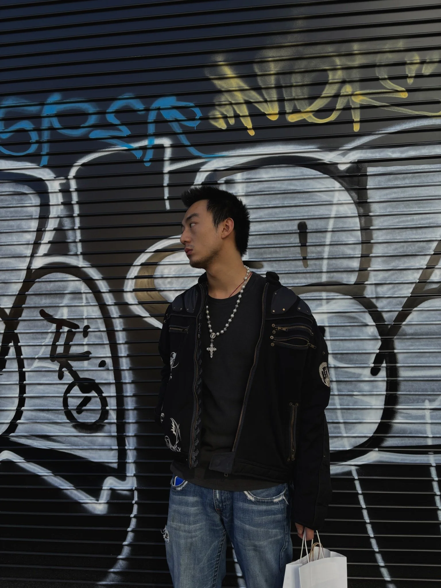 A young man with short dark hair, wearing a black jacket with white embroidery and a pearl necklace, standing in front of a black metal wall with graffiti, holding shopping bags in his right hand.