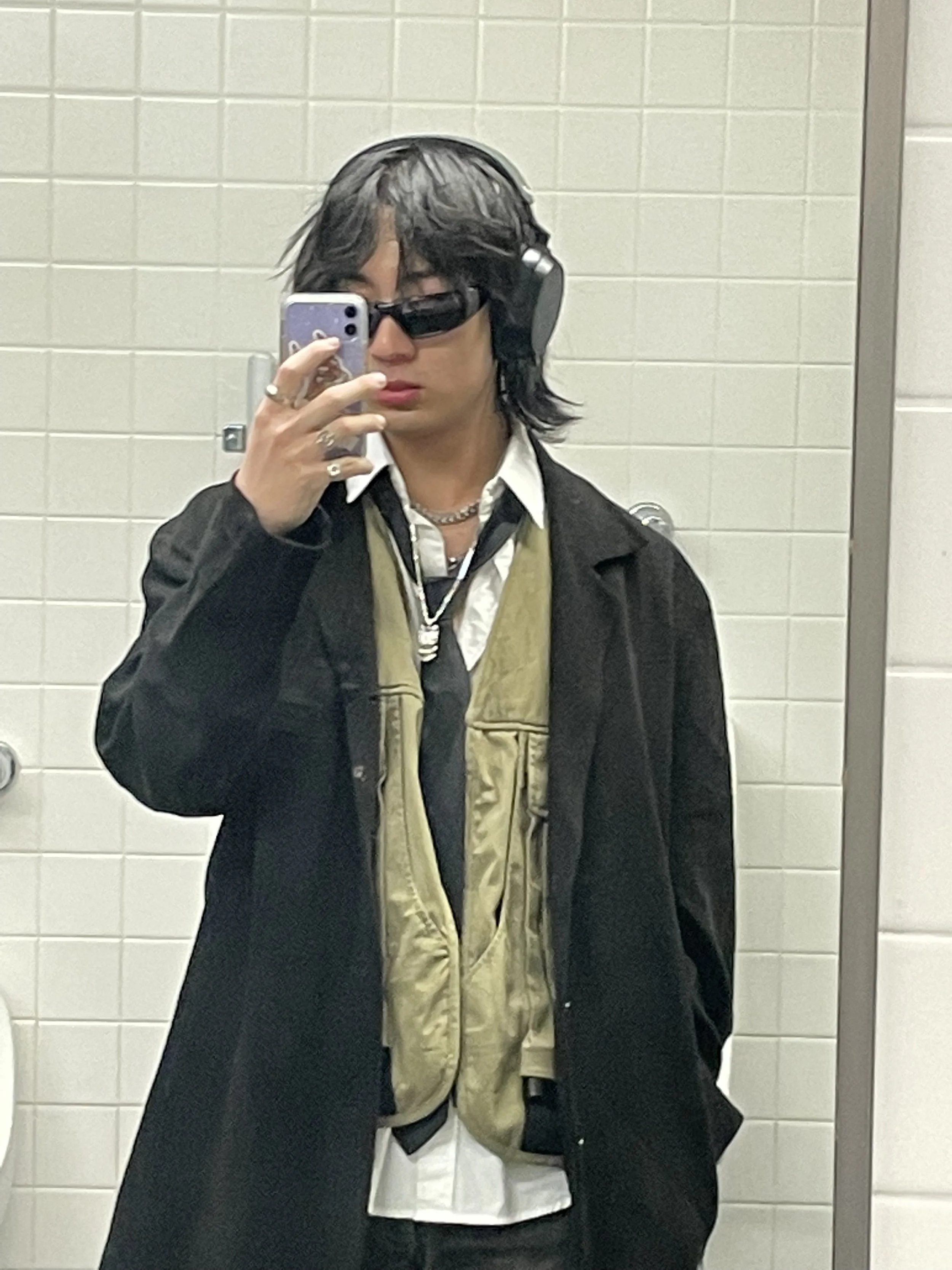 tu. taking a mirror selfie in a bathroom, wearing dark sunglasses, headphones, a white collared shirt, a beige vest, and a black jacket.