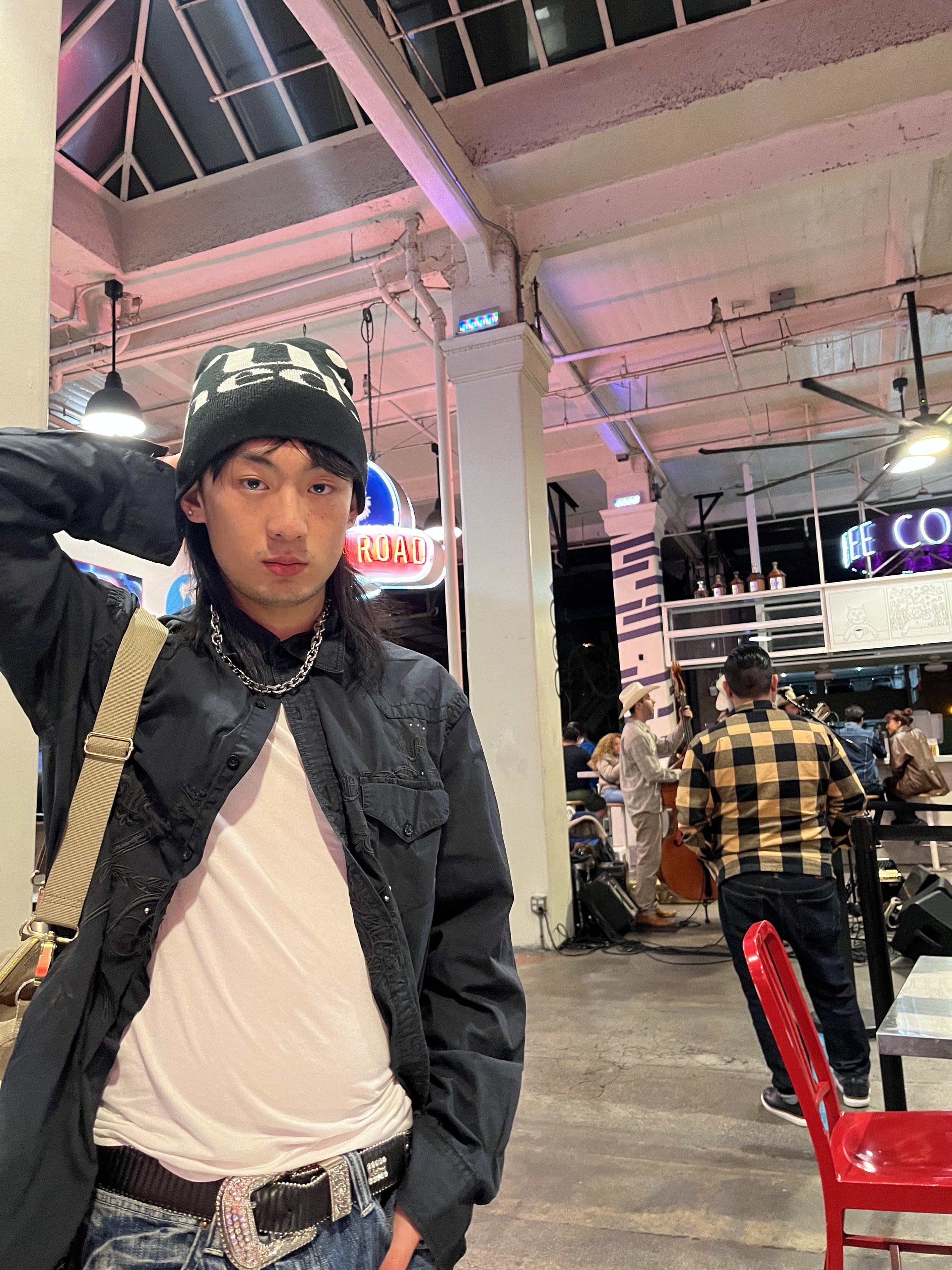 A young man with long dark hair, wearing a black beanie with a white pattern, a black jacket, a white shirt, and a silver chain necklace, standing in a busy indoor space with people sitting and standing in the background, including a woman in a cowbo