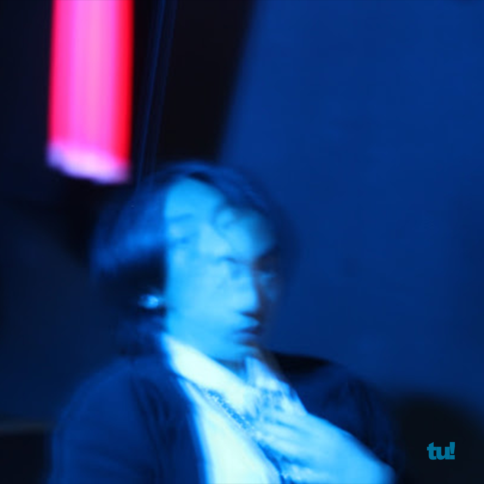 Close-up of a person's face illuminated by blue light, with dark hair and a serious expression, wearing a jacket, in a dimly lit setting.