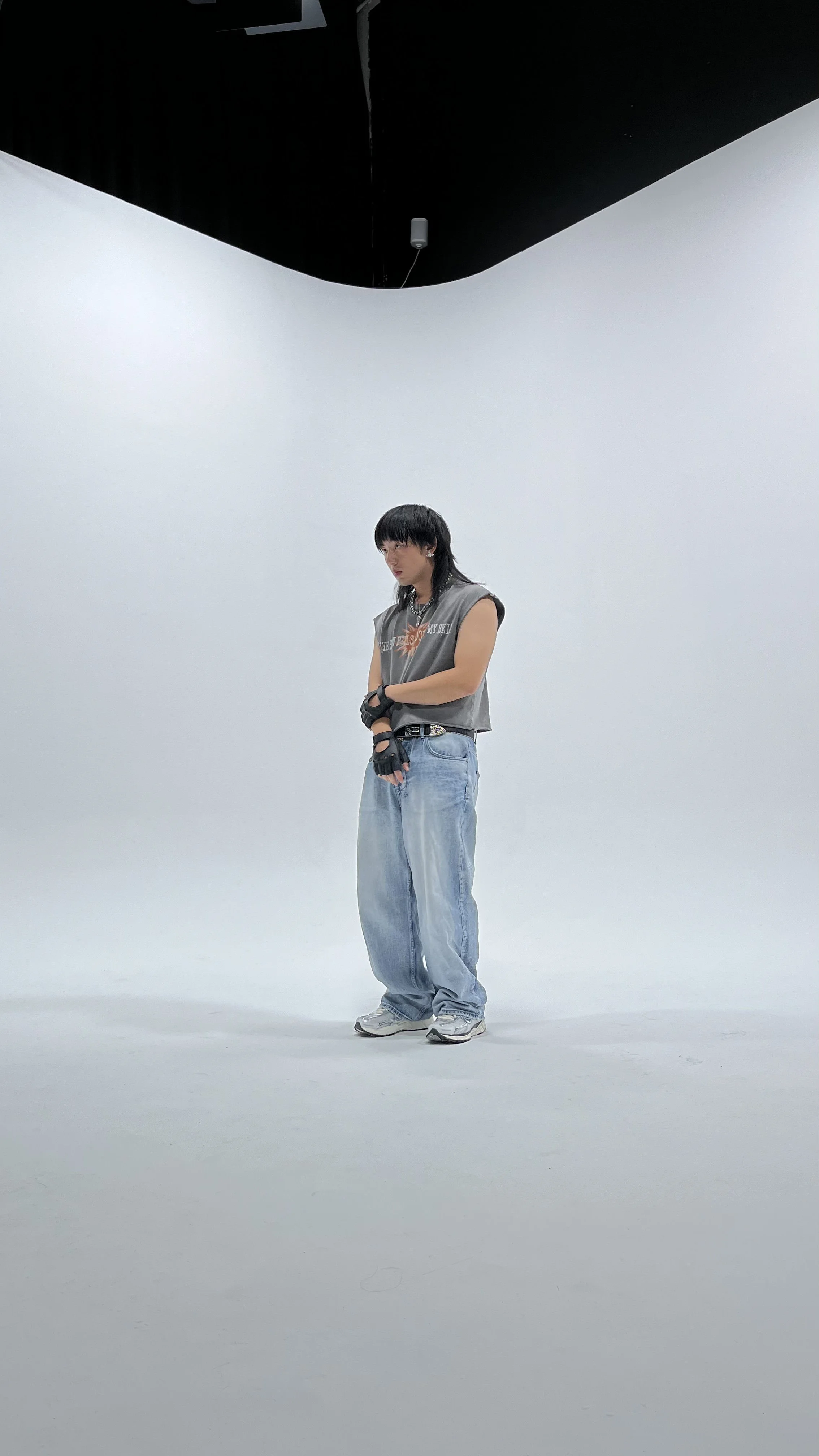 A person with dark hair and bangs, wearing a sleeveless top, baggy jeans, sneakers, and gloves, standing in a white cyc wall studio with black ceiling panels.
