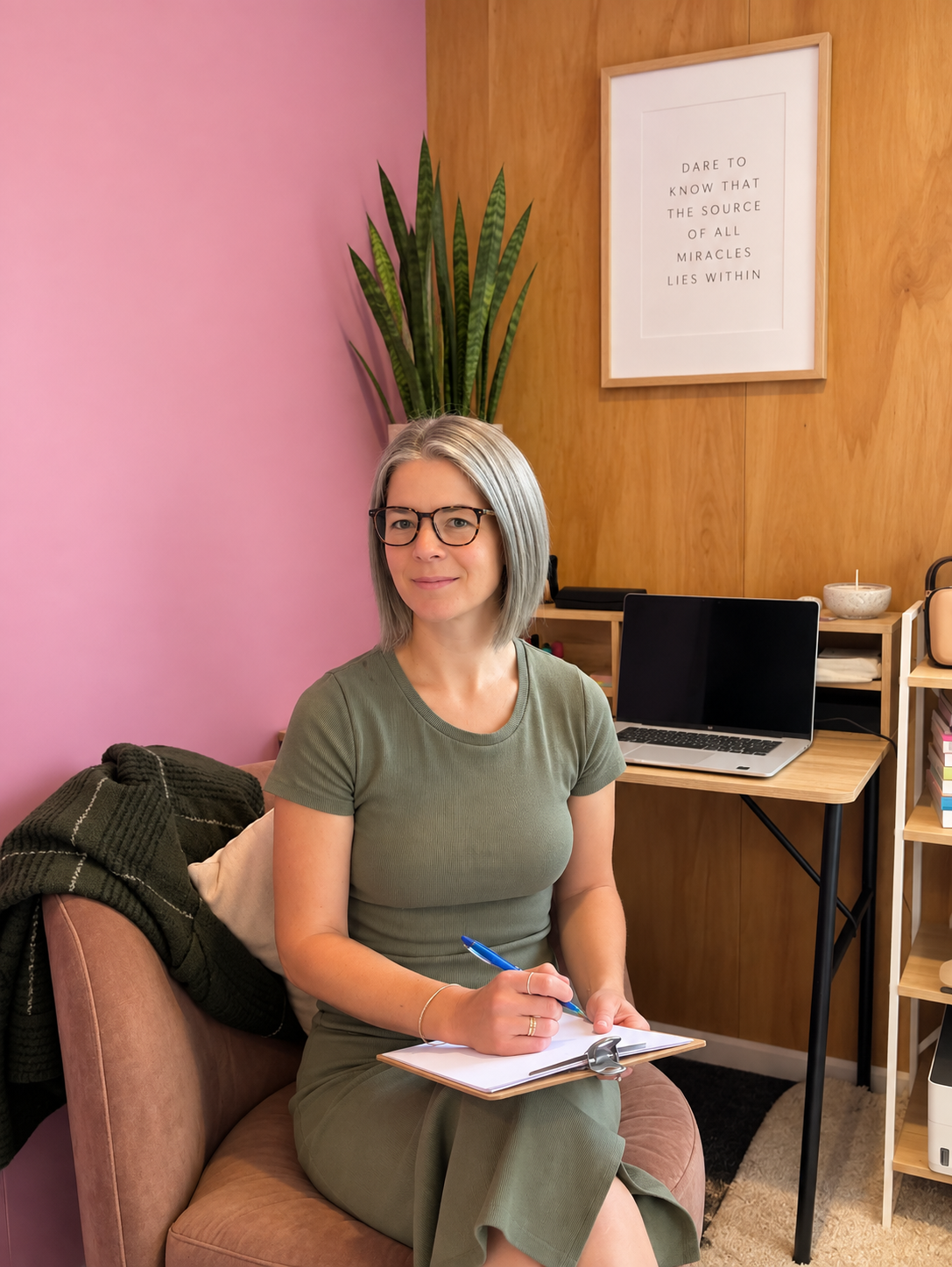A woman with gray hair and glasses sitting on a pink couch, writing on a clipboard, in a room with a pink wall, a wooden wall, a large plant, a framed quote, a laptop, and shelves with books and decor.