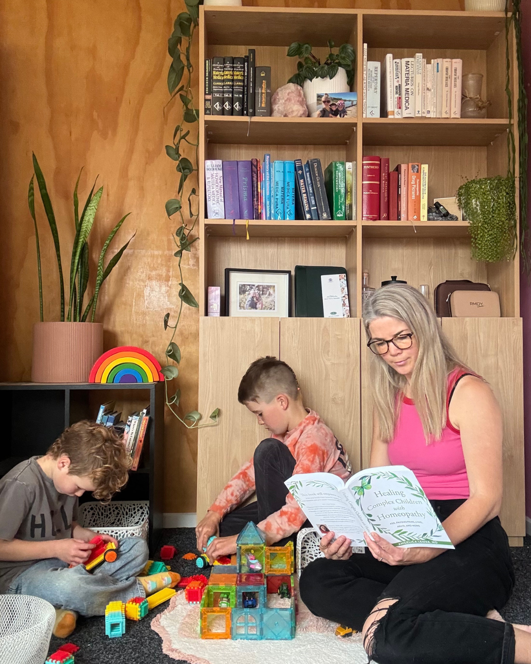 A woman and two young boys playing with colorful building blocks on the carpeted floor in a cozy room with wooden shelves filled with books, plants, and decorative items.