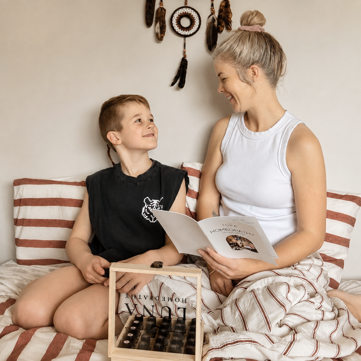 A woman and a boy sitting on a bed, smiling and looking at each other. The woman is holding a booklet titled "Homemopathy" and the boy is holding a wooden box with essential oils. Backdrop includes a dreamcatcher decor on the wall.