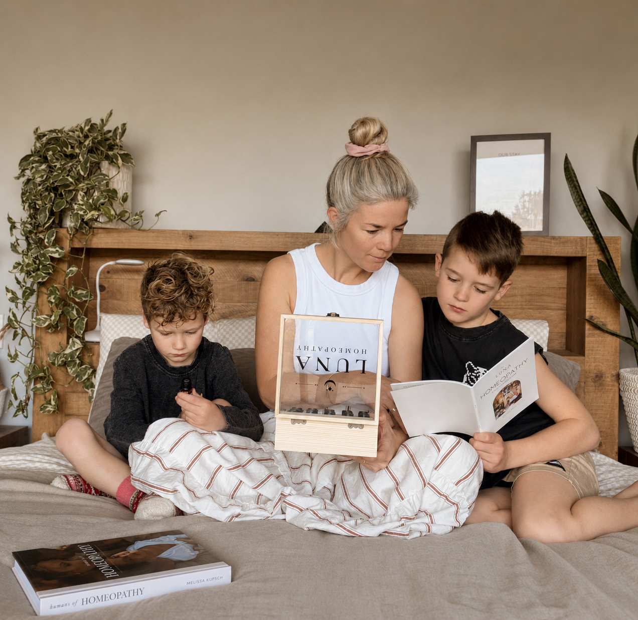 A woman and two children sitting on a bed, reading books together in a cozy bedroom with a wooden headboard, plants, and natural light.