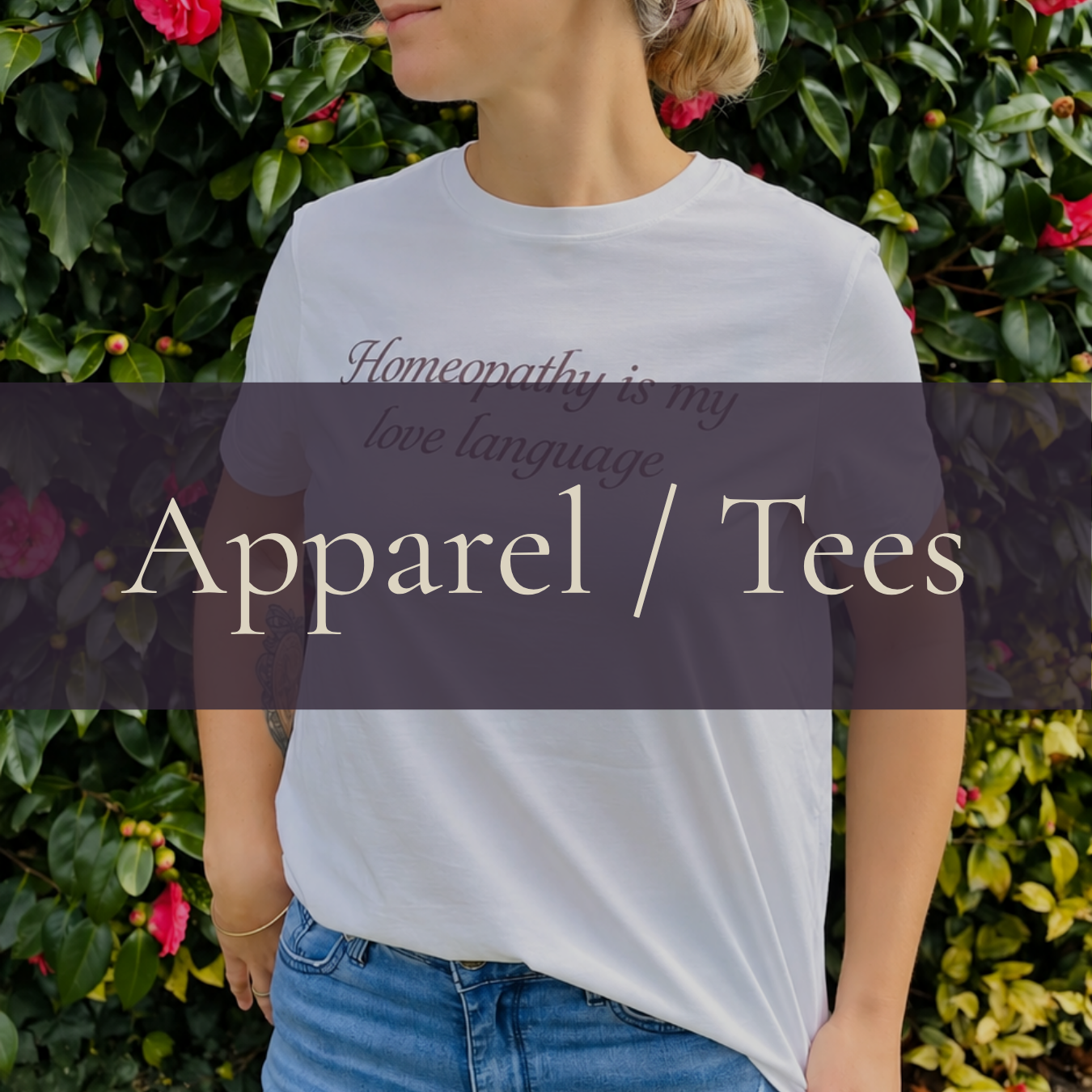 A person wearing a white T-shirt with the text 'Homeopathy is my love language' standing in front of greenery with pink flowers. The overlay text says 'Apparel / Tees'.