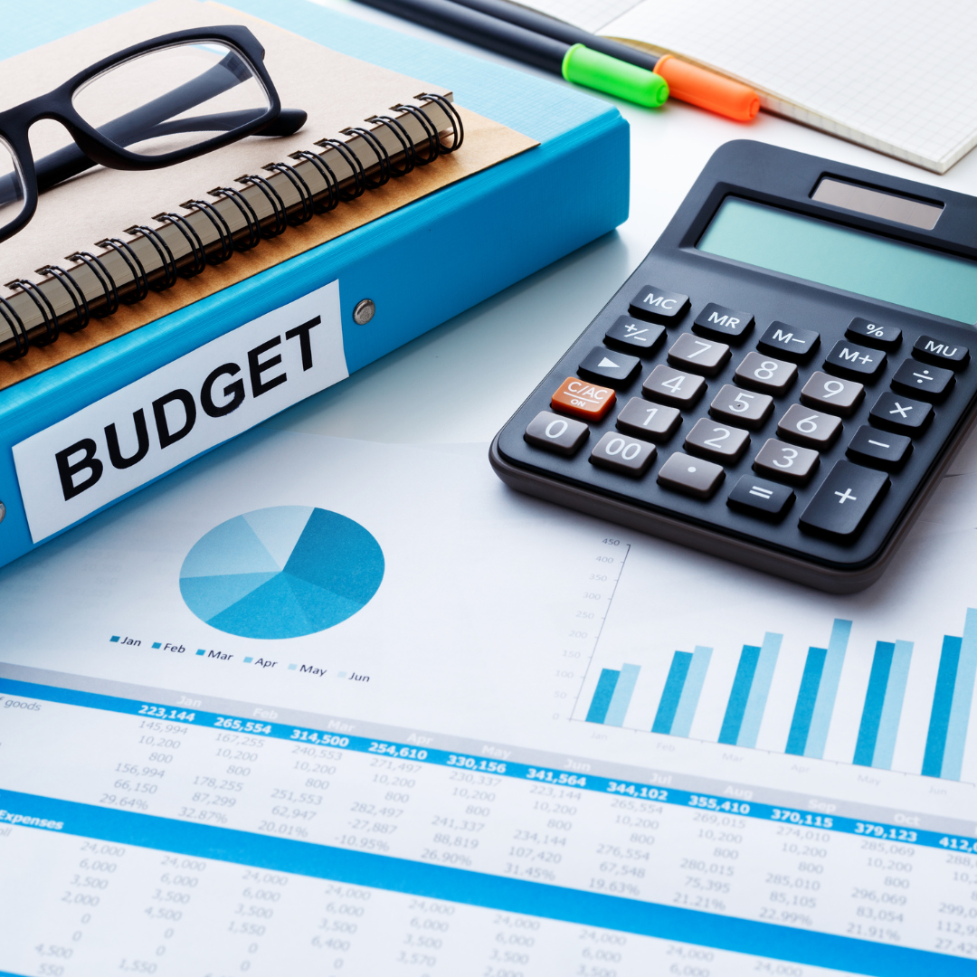 Office desk with a budget binder labeled 'BUDGET', a calculator, a pie chart on paper, a pair of eyeglasses, markers, and notebooks.