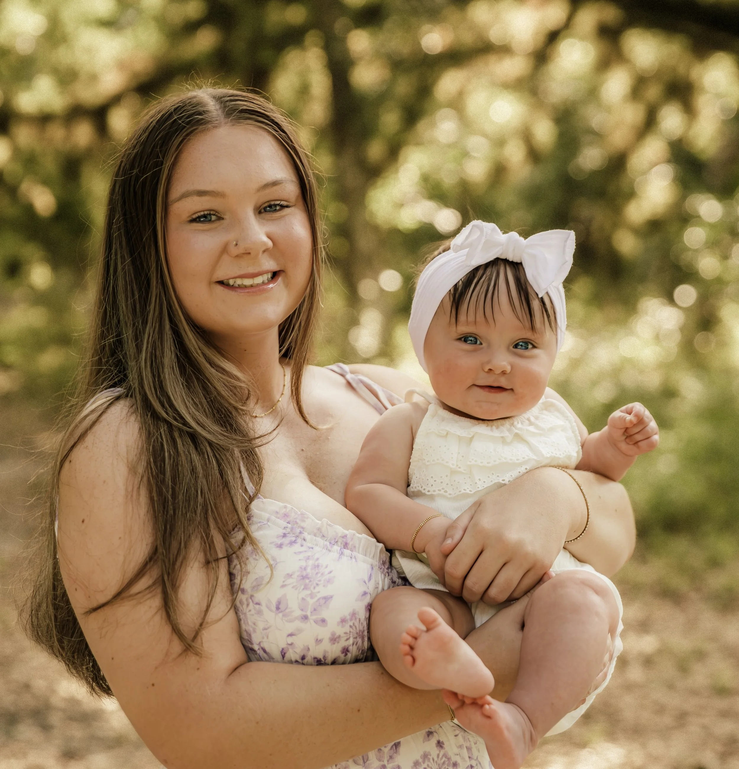 A woman smiling while holding a baby girl outside, in front of trees with sunlight creating a bokeh effect in the background.