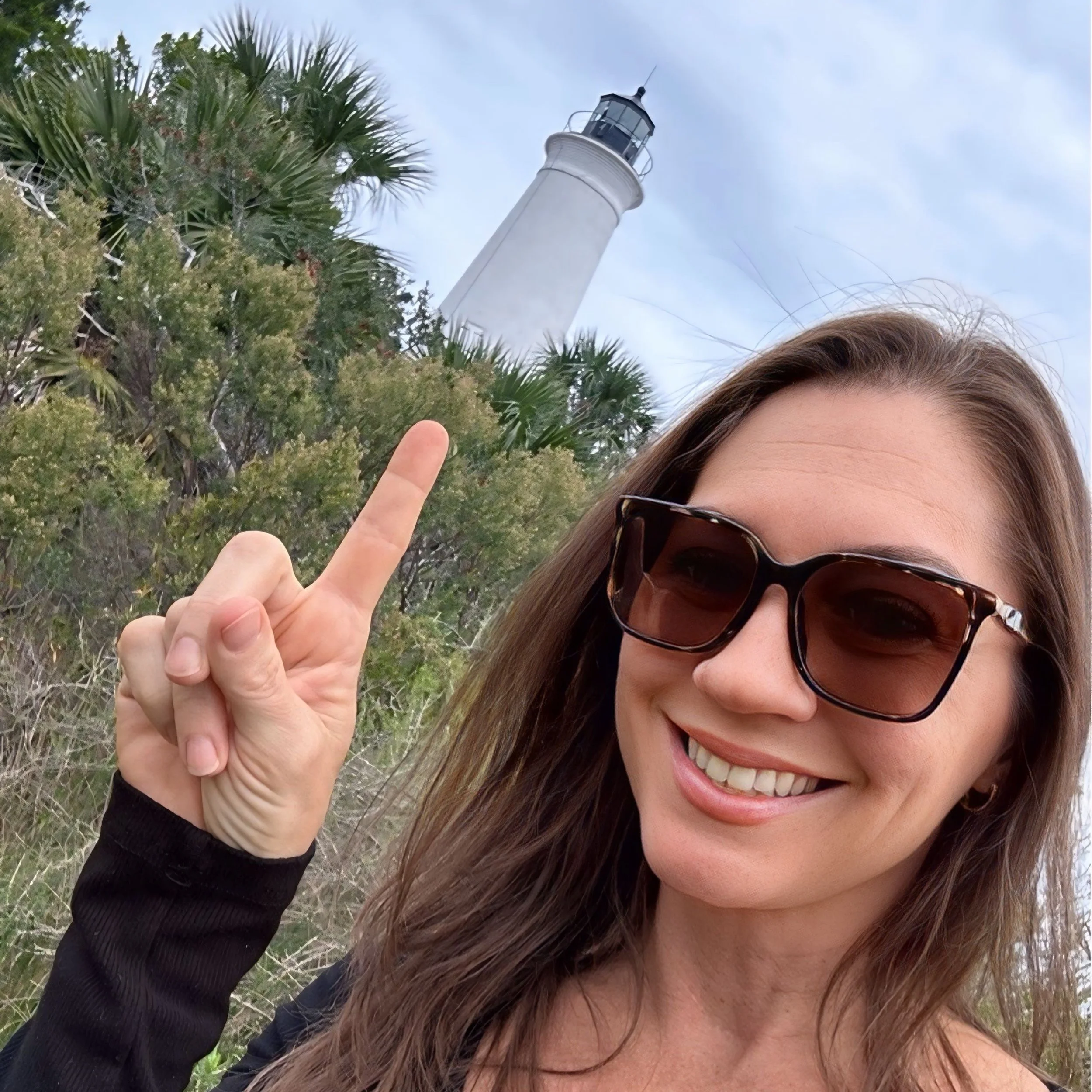A woman with sunglasses and long brown hair smiling outdoors, showing her teeth. She is holding up her hand with two fingers in a peace sign. Behind her are green trees, and a white lighthouse is visible in the sky.