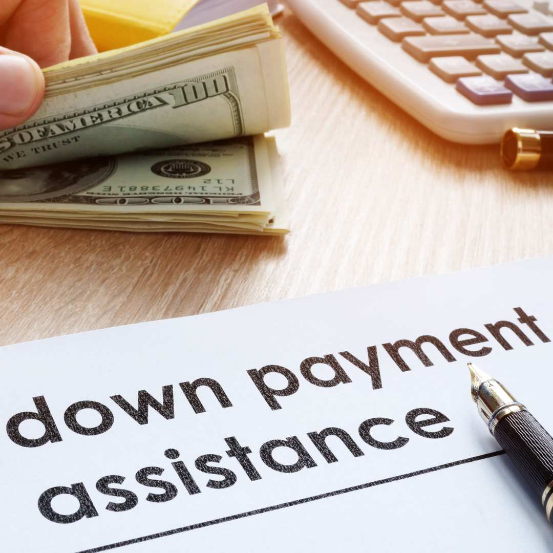 A stack of hundred-dollar bills, a calculator, and a pen on a wooden desk, with a document titled 'down payment assistance' in the foreground.