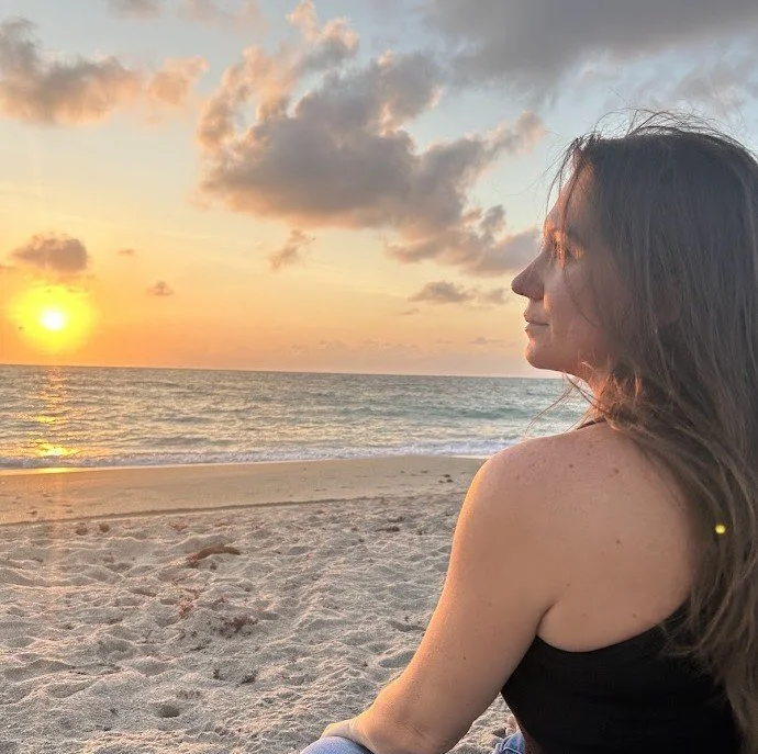 A woman sitting on the beach watching the sunset over the ocean, with clouds in the sky and the sun near the horizon.