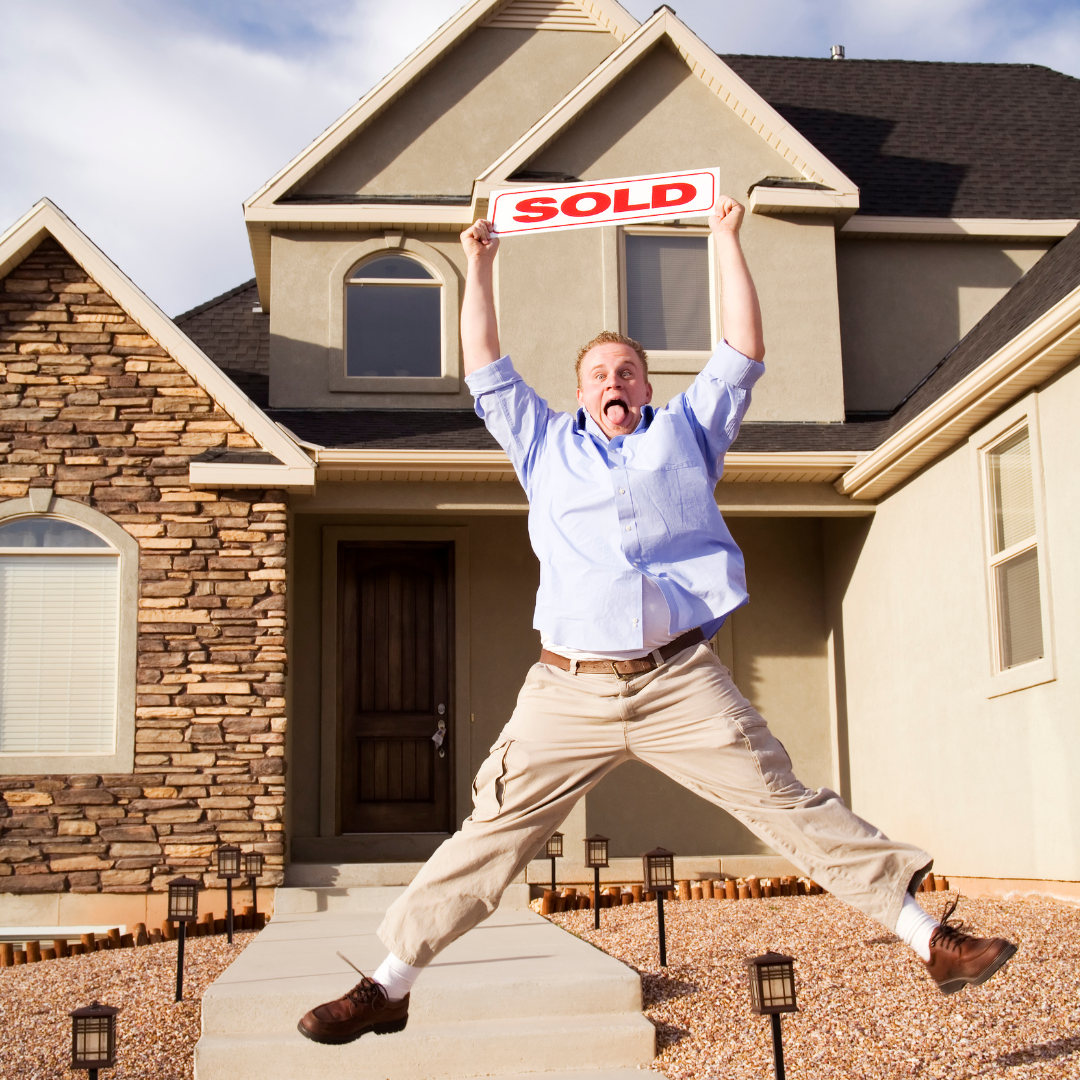A excited man in a blue shirt and beige pants jumping in front of a house holding a "SOLD" sign, indicating the house has been purchased.