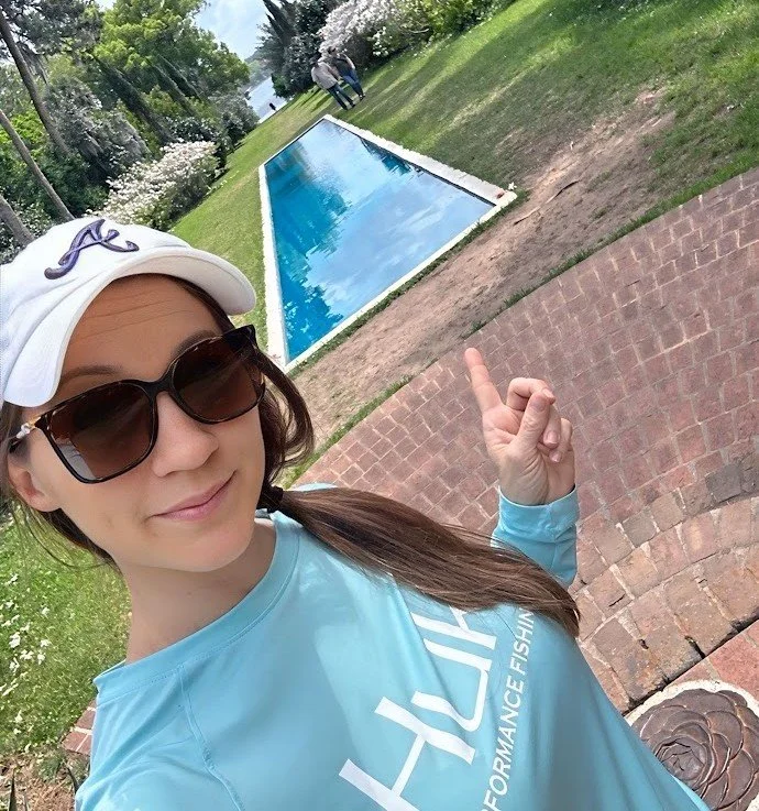 A woman wearing sunglasses and a white cap with an 'A' logo, pointing at a swimming pool outdoors, with a grassy area and trees in the background.