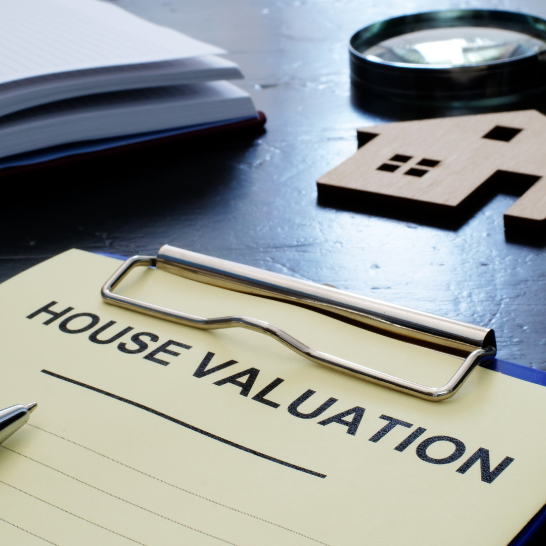 A clipboard with a house valuation form on a dark table, along with a small wooden house cutout, a magnifying glass, and a pen.