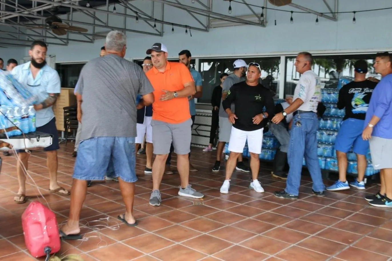 A group of men are gathered in an outdoor covered area, some are talking and others are waiting near a stack of bottled water. The setting appears casual and possibly related to a distribution or outdoor event.