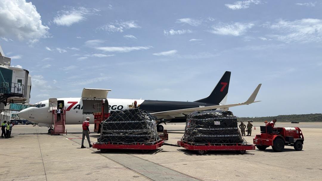 Cargo plane on tarmac with cargo containers being loaded, ground crew and personnel nearby, clear sky.